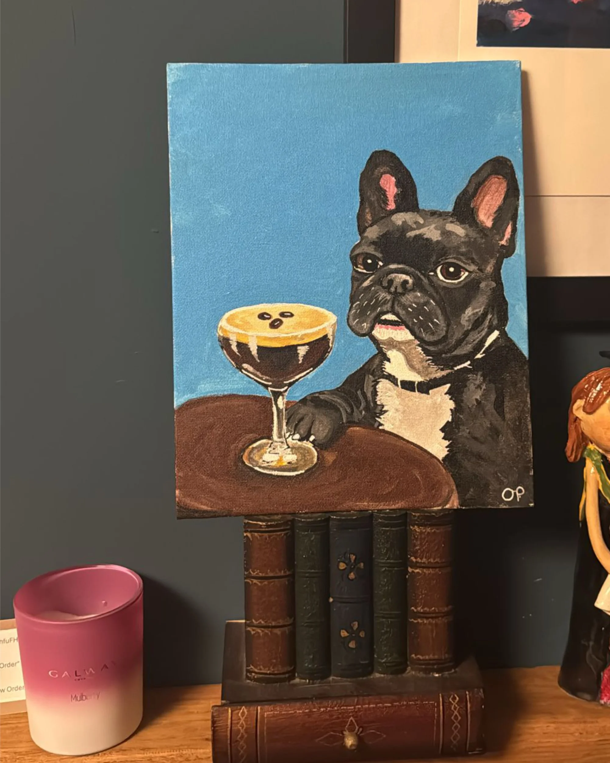 Painting of a French Bulldog with a black coat, sitting at a table with a glass of dark beverage topped with foam and coffee beans. The background is blue with a darker blue foreground; artwork is on a small bookshelf-shaped stand.