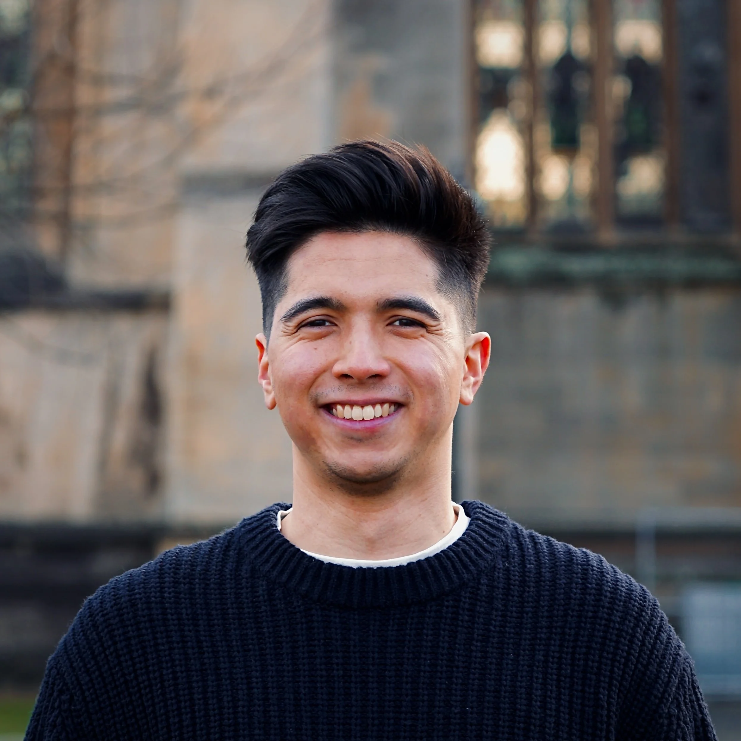 A smiling young man with dark, styled hair wearing a black sweater outdoors, with a blurred background of trees and a building.