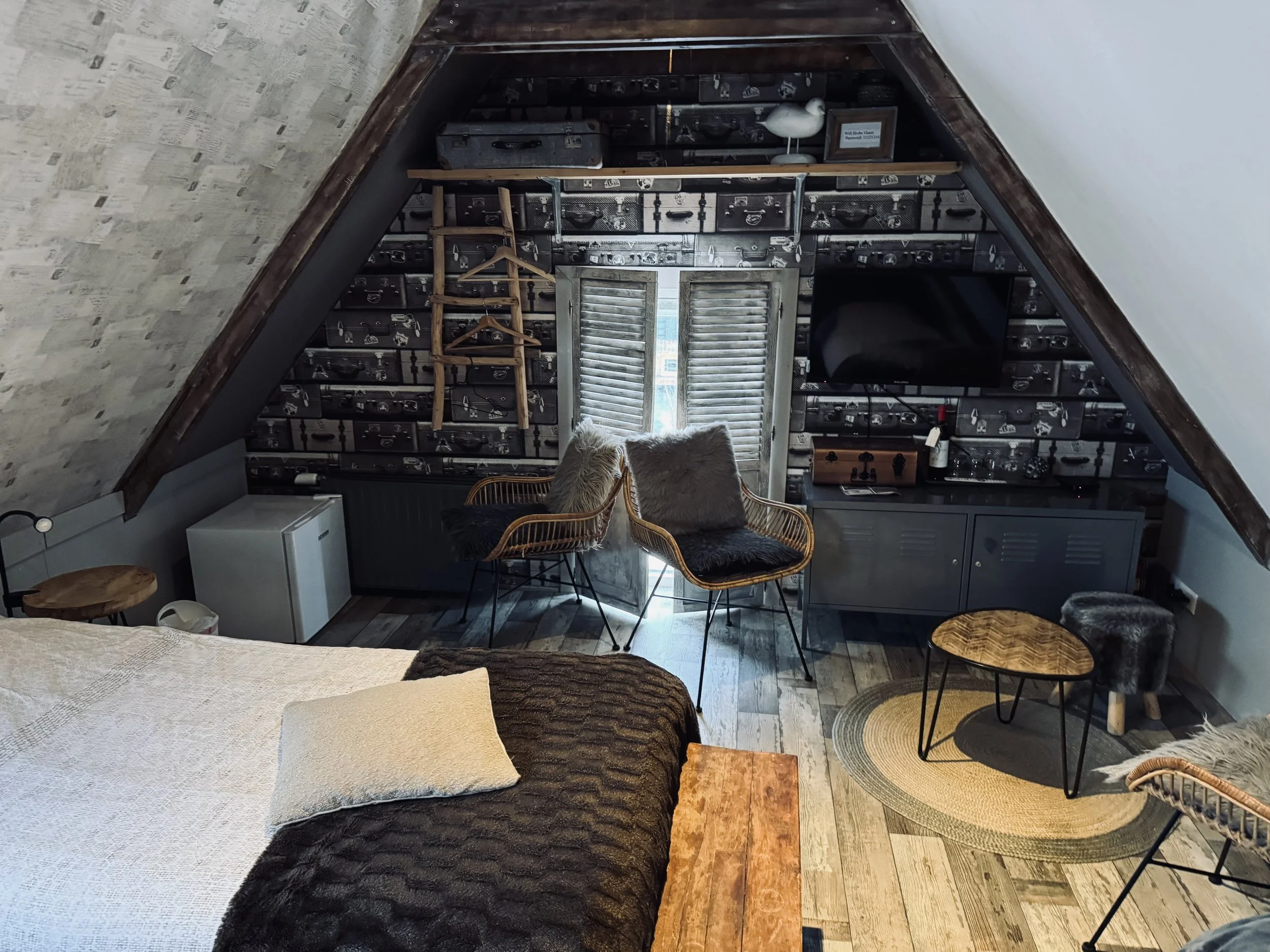 Gezellige zolderkamer met schuin plafond, houten vloer, bed met zwart-wit beddengoed en een zithoek met twee stoelen, een kleine ronde tafel en een vloerkleed, met een muur met zwart-wit patroon behang