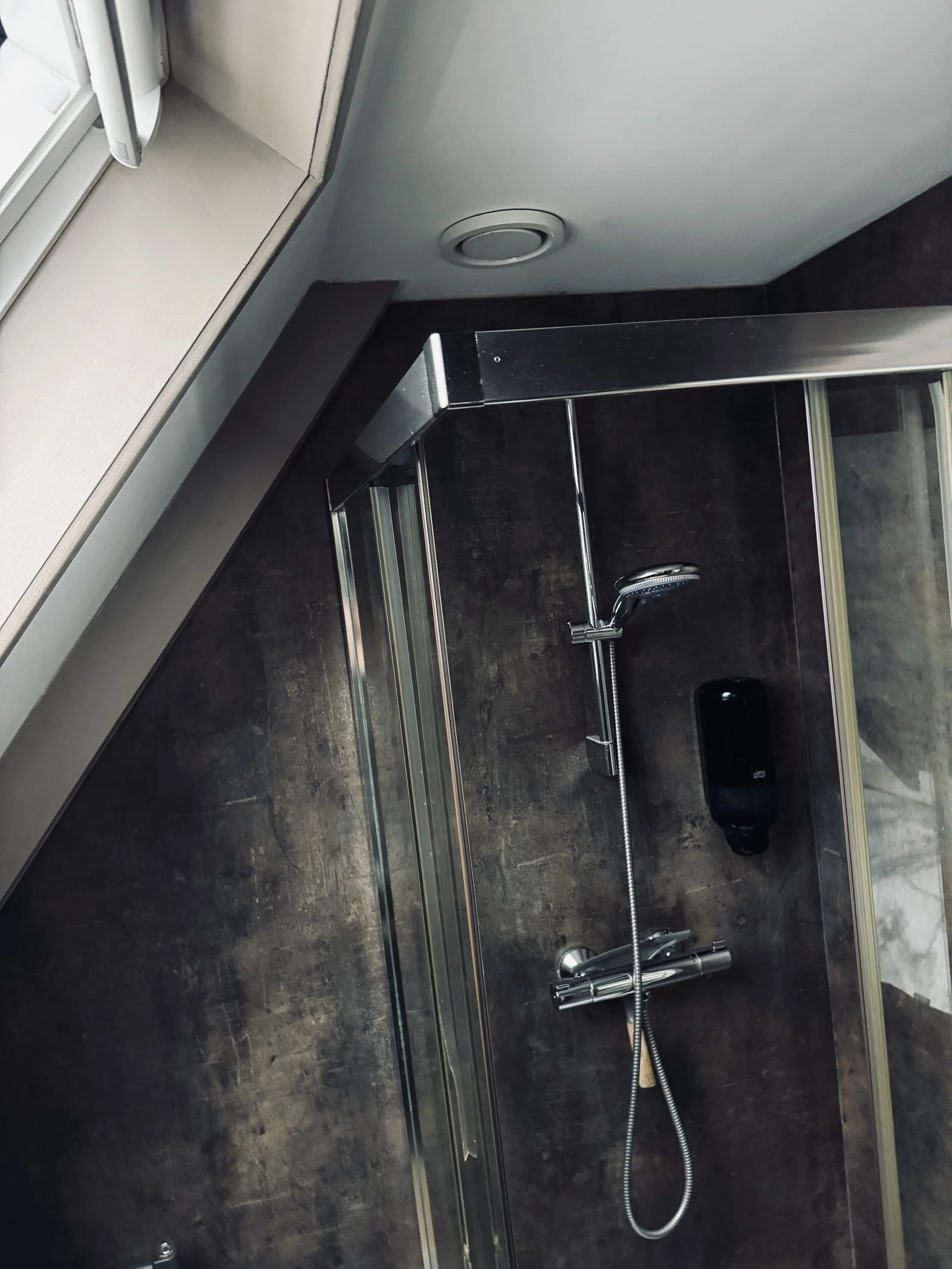 Een moderne douche met donkerbruine tegelwanden, zilveren armaturen, waaronder een handdouche, en een zwarte zeepdispenser aan de muur.