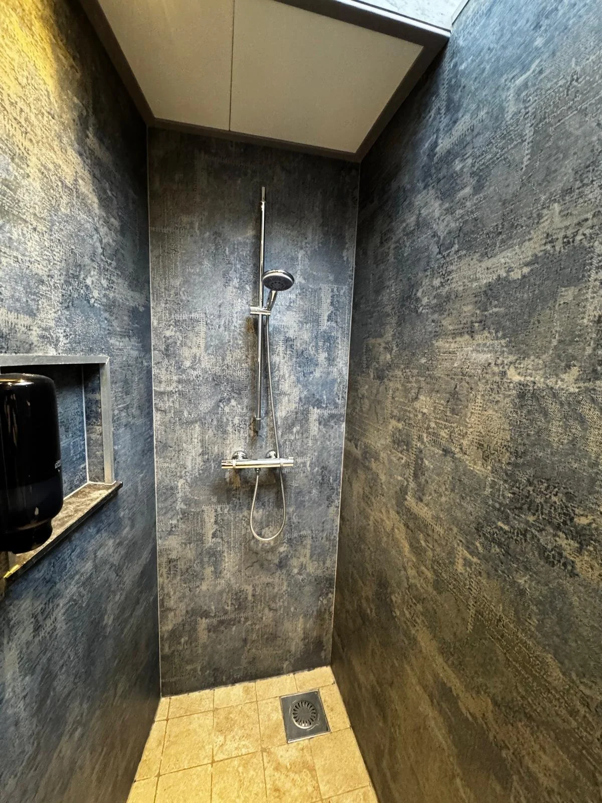 A small, modern walk-in shower with dark textured wall tiles, a chrome showerhead, and a beige tiled floor with a floor drain.