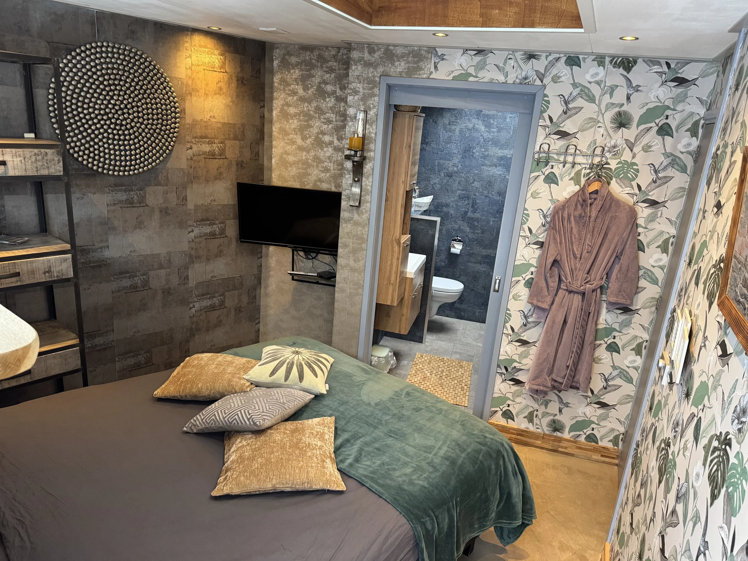 Een gezellige slaapkamer met een bed versierd met sierkussens, een aan de muur bevestigde flatscreen-tv, een gestructureerde muur en een badkamer die zichtbaar is door een open deur. Een roze badjas hangt aan een haak aan een muur uniek behang