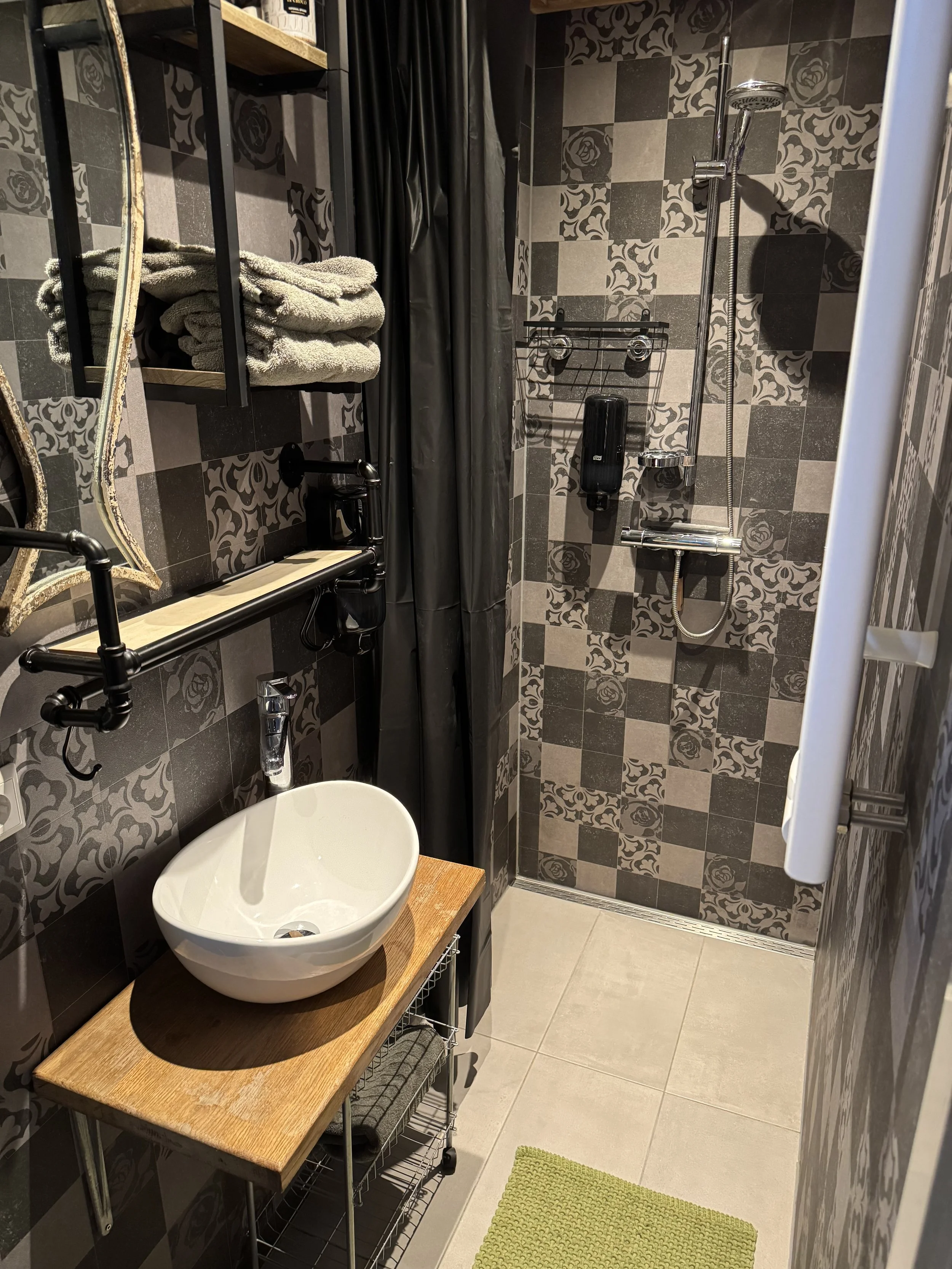 Kleine badkamer met zwarte en grijze wandtegels met patroon, een inloopdouche met handdouche, een wandgemonteerde zeepdispenser en een zwart douchegordijn. Op een houten plank liggen opgevouwen grijze handdoeken boven een spiegel met een zwarte lijst