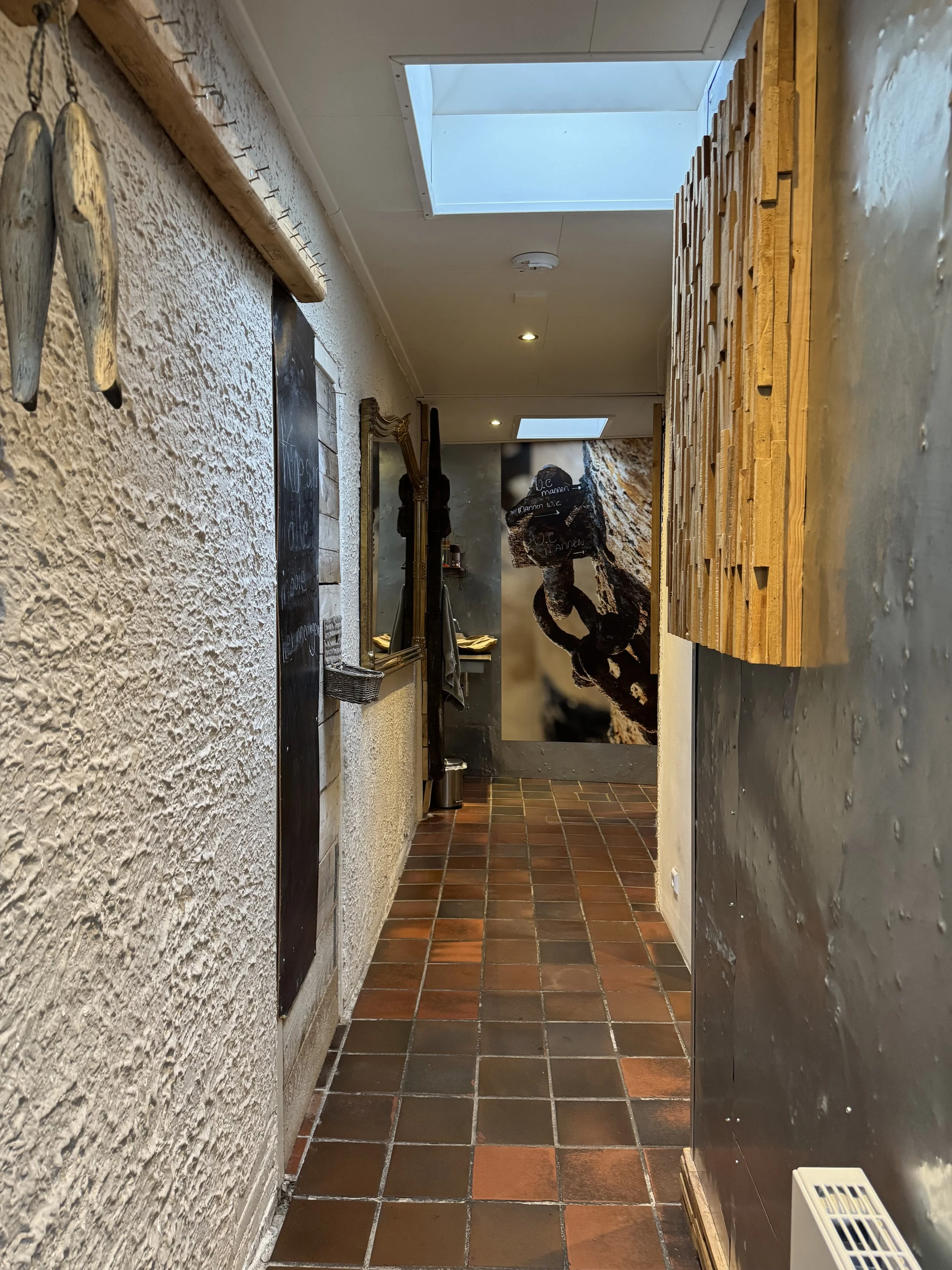 Gezellige hal met rustieke inrichting, waaronder een gestructureerde muur, een spiegel en een grote muurschildering van iets. Het plafond heeft dakramen en inbouwverlichting, met een houten accentmuur aan de rechterkant.
