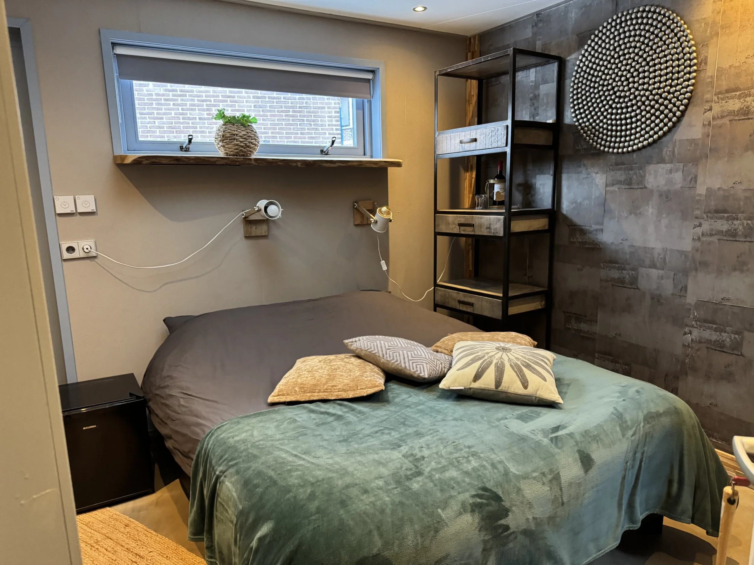Een moderne slaapkamer met een bed bedekt met een groene fluwelen deken en decoratieve kussens, een kleine zwarte koelkast naast het bed, een raam met een potplant, aan de muur bevestigde leeslampjes, een zwarte metalen en houten rek