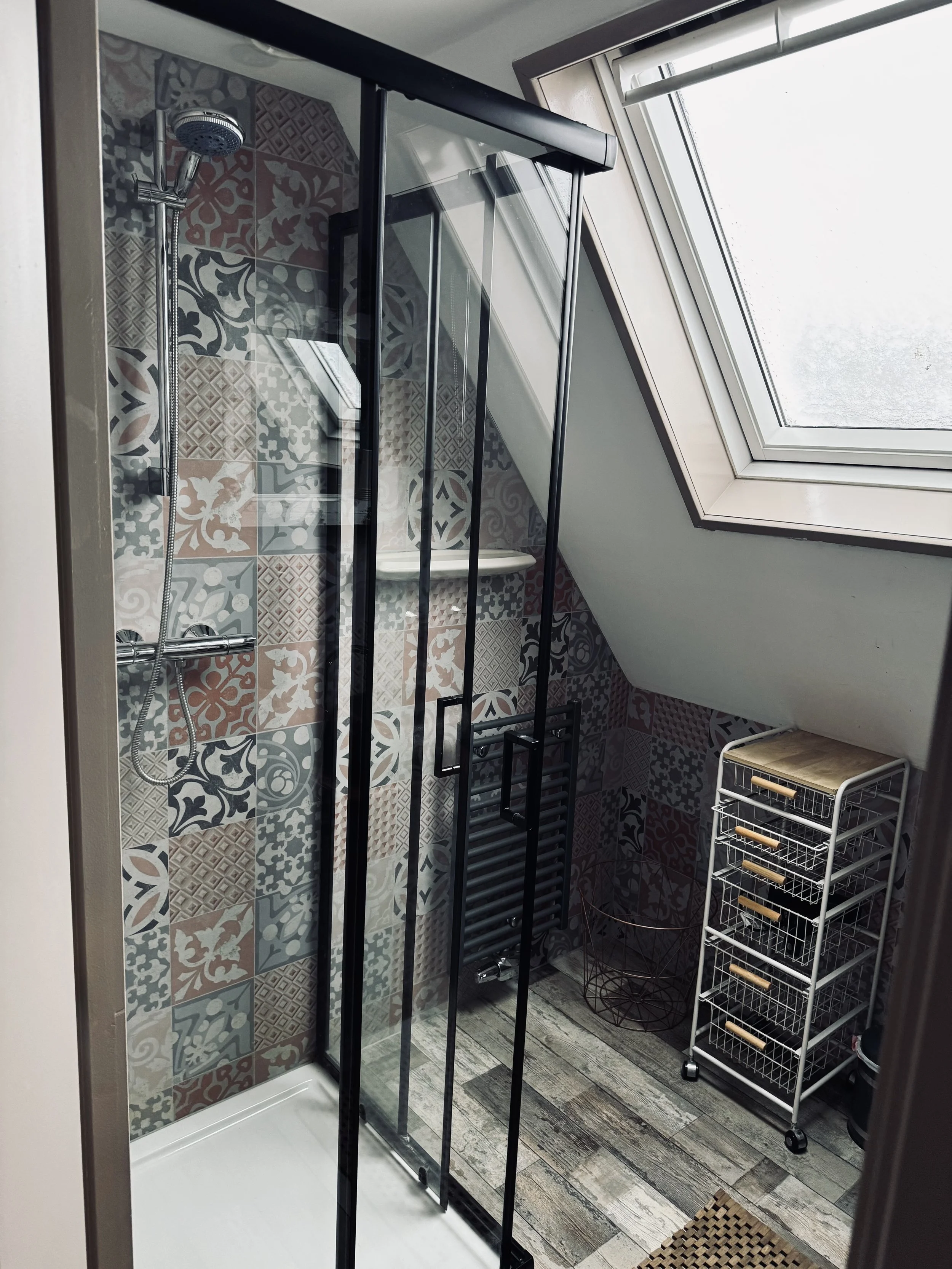 Badkamer met een hoekdouchecabine, patroontegels op de muur, een dakraam en een wit opbergrek.