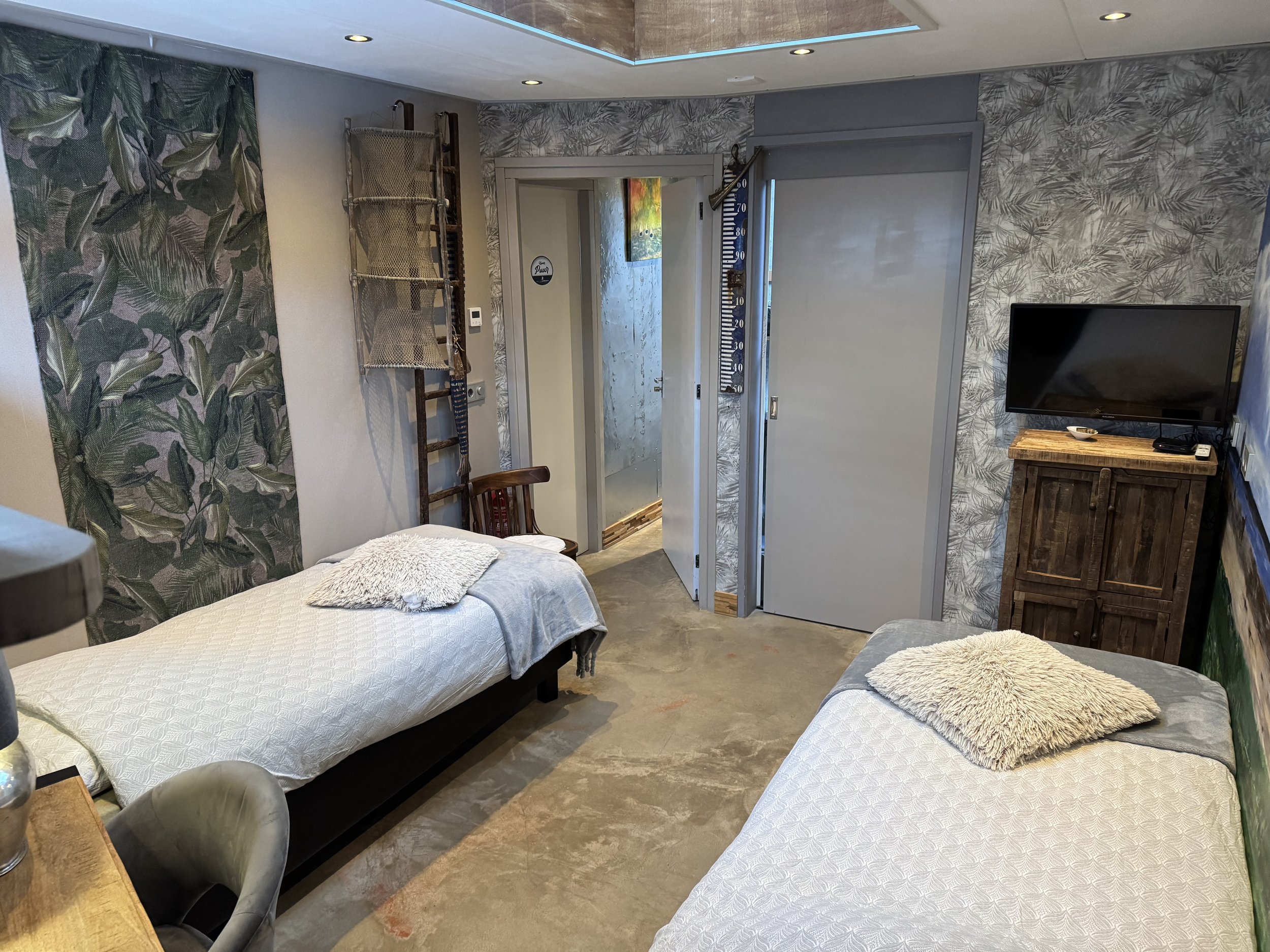 Een kamer met twee eenpersoonsbedden bedekt met witte dekbedden en zachte kussens, een flatscreen-tv op een houten kast, een houten stoel en decoratief behang met tropisch thema.