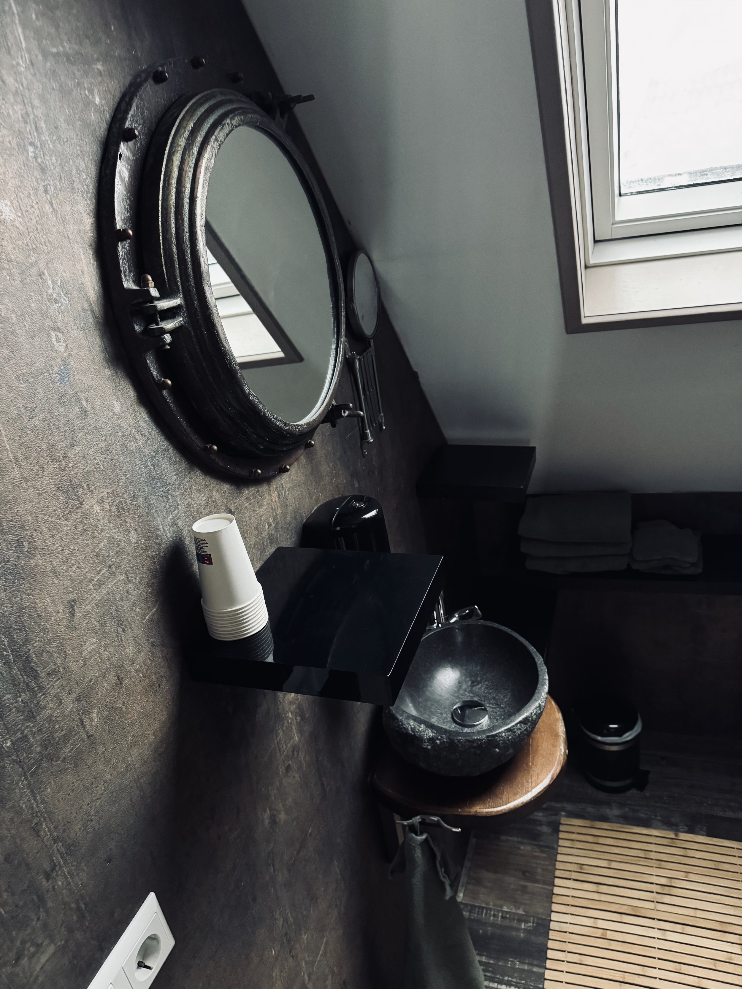 Badkamer met een ronde spiegel met zwarte lijst, stenen wastafel op houten onderstel, raam, opgevouwen handdoeken, kopjes en toiletartikelen.