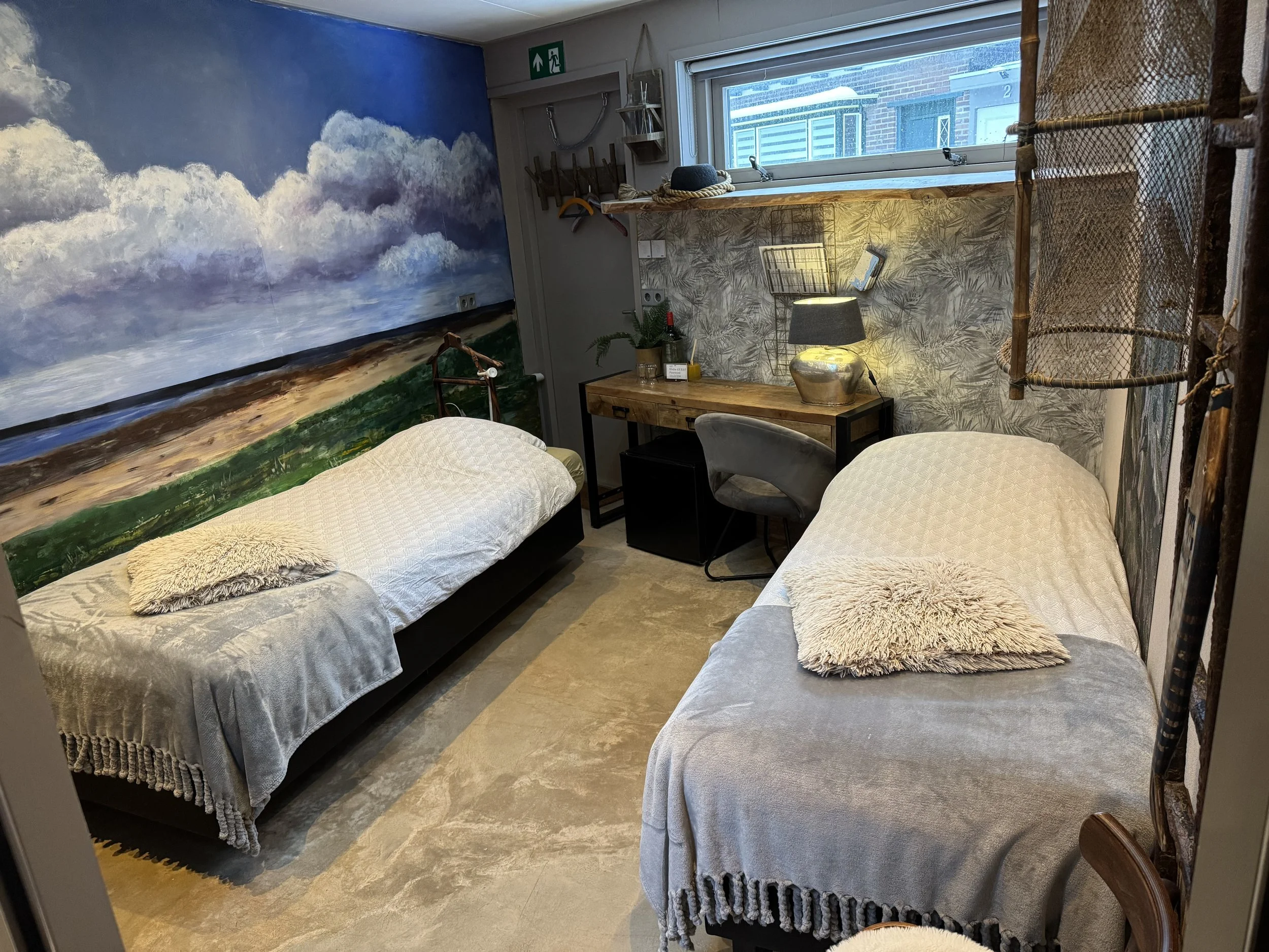 Een gezellige kamer met twee eenpersoonsbedden, één aan elke kant, met witte dekens en zachte kussens, een klein bureau met een lamp, een raam met uitzicht op de naburige gebouwen en een grote wolkenmuurschildering op de muur achter één van de bedden