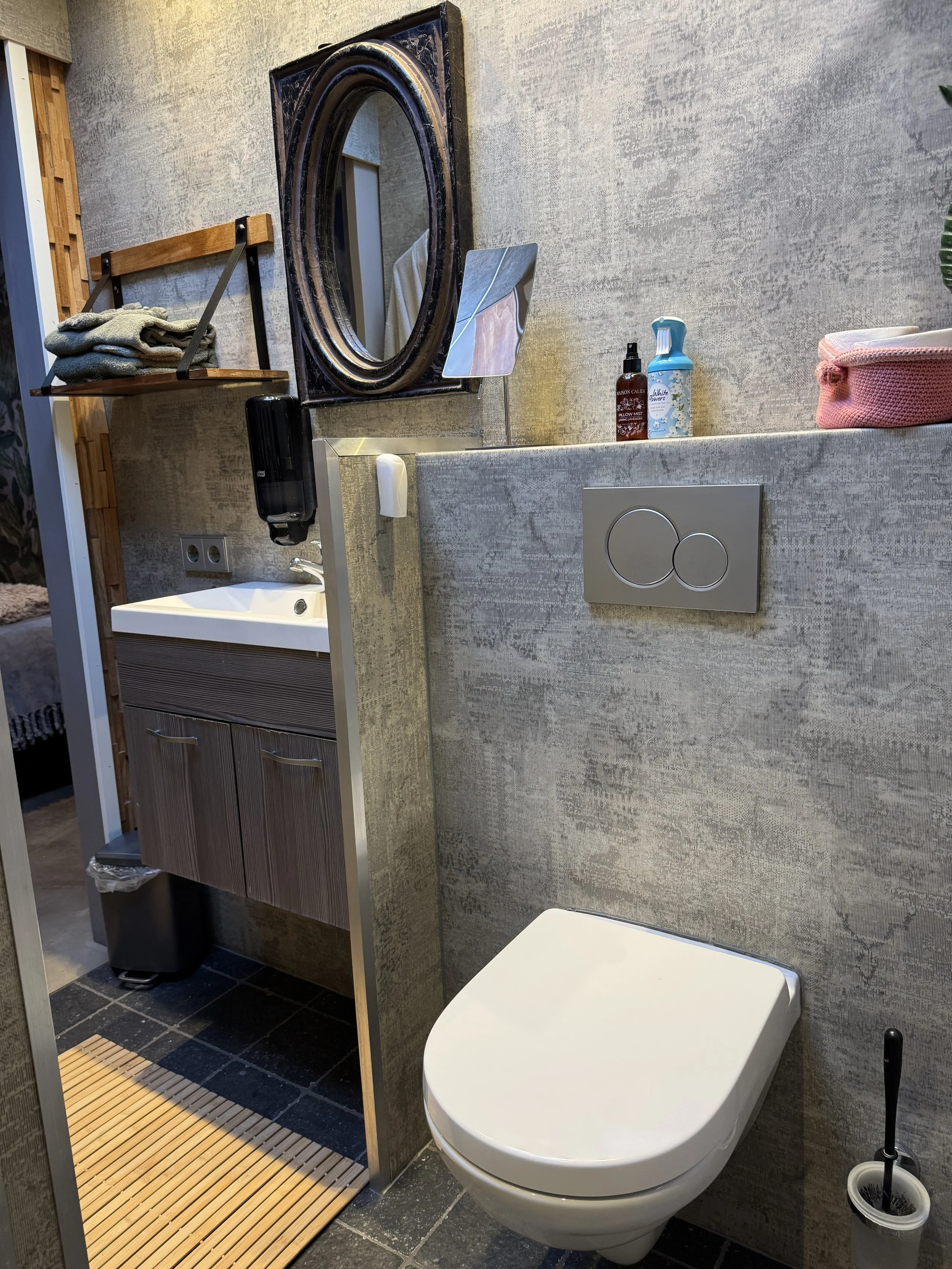 Een compacte badkamer met een modern wandtoilet, een kleine wastafel met spiegel en planken met handdoeken, zeep en schoonmaakspullen. De muren zijn bekleed met structuurbehang en de vloer is betegeld met donkere stenen tegels.