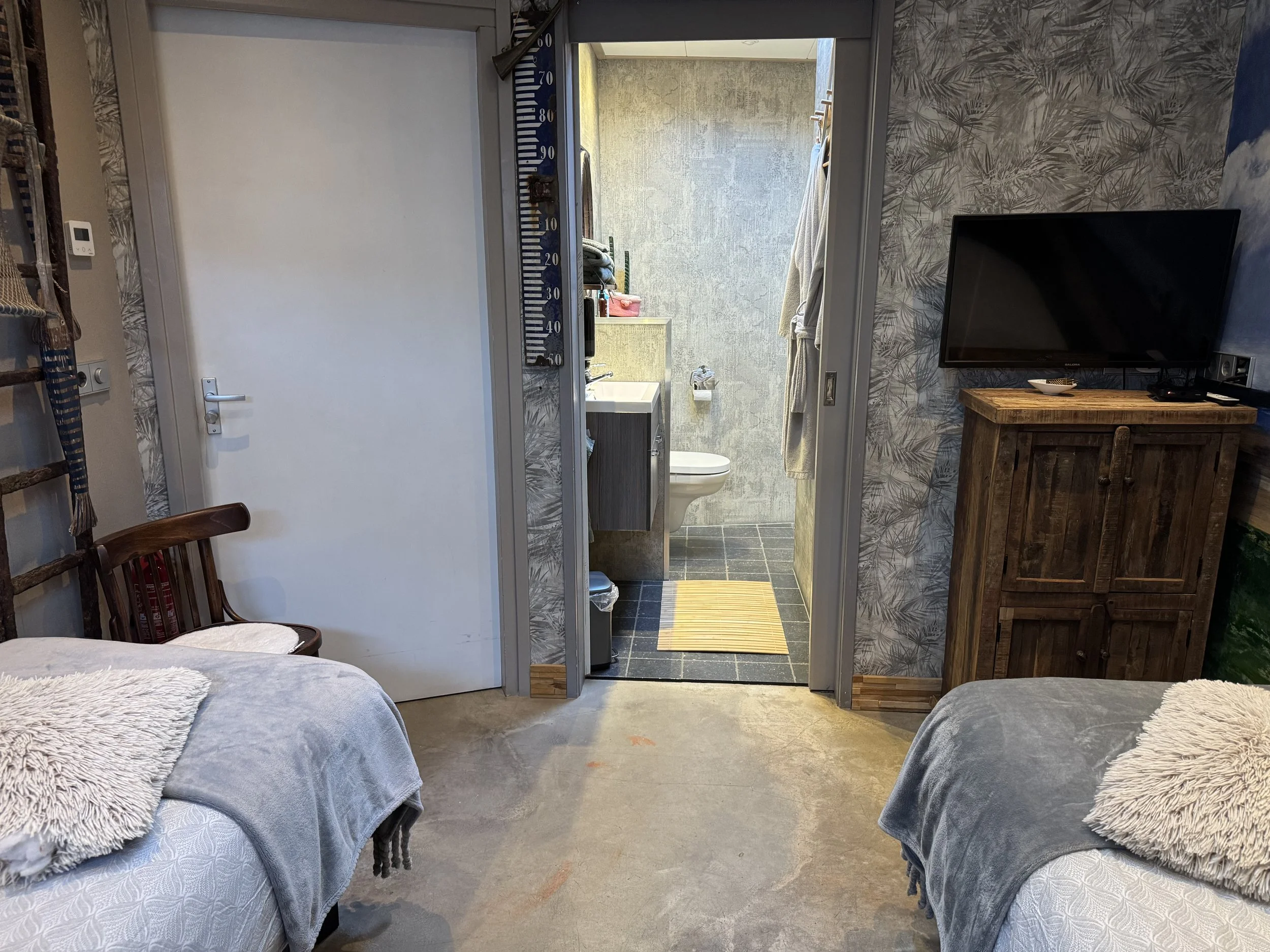 Uitzicht op een gezellige slaapkamer met twee bedden, een tv op een houten kast en een open deur die naar een kleine badkamer met toilet en wastafel leidt.