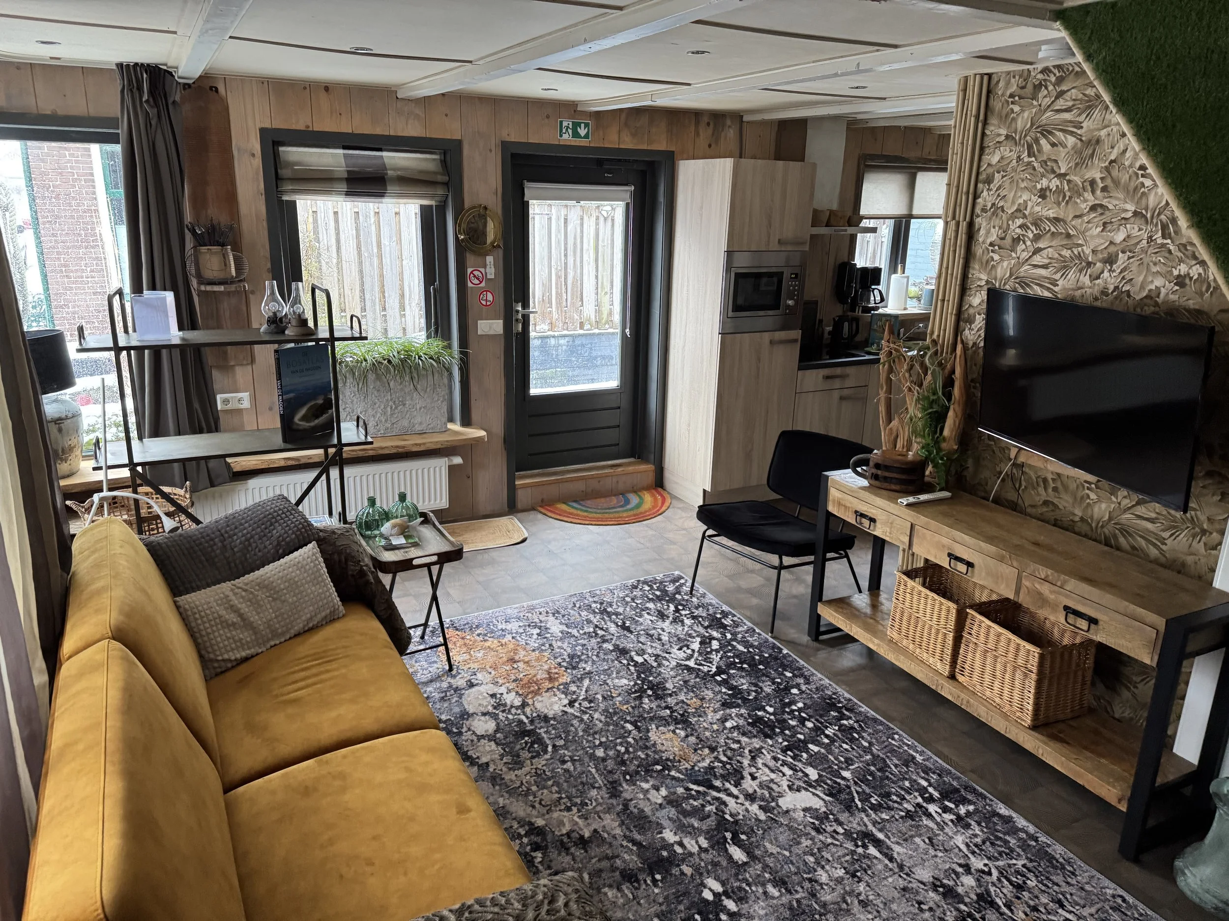 Een gezellige woonkamer met een mosterdkleurige bank, een zwarte stoel, een tv aan de muur en een rustieke houten entertainmentconsole met manden. De kamer heeft houten wanden, behang met patronen, een groot raam met bruine gordijnen en diverse decor