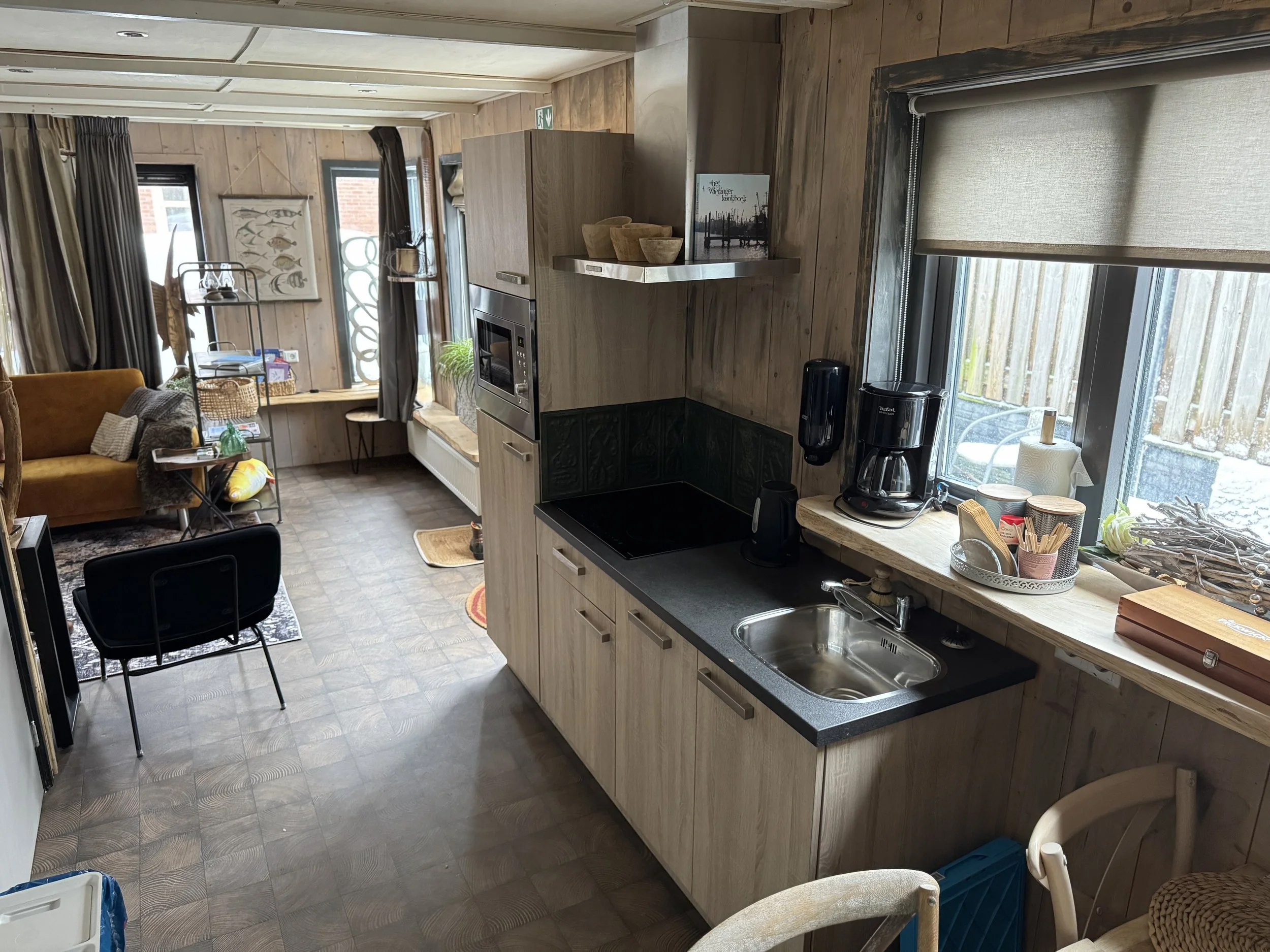 Keuken met zwart aanrechtblad, roestvrijstalen spoelbak, koffiezetapparaat, magnetron, raam met beige rolgordijn, houten kasten en woonkamer met bank, stoel en kunst aan de muur.