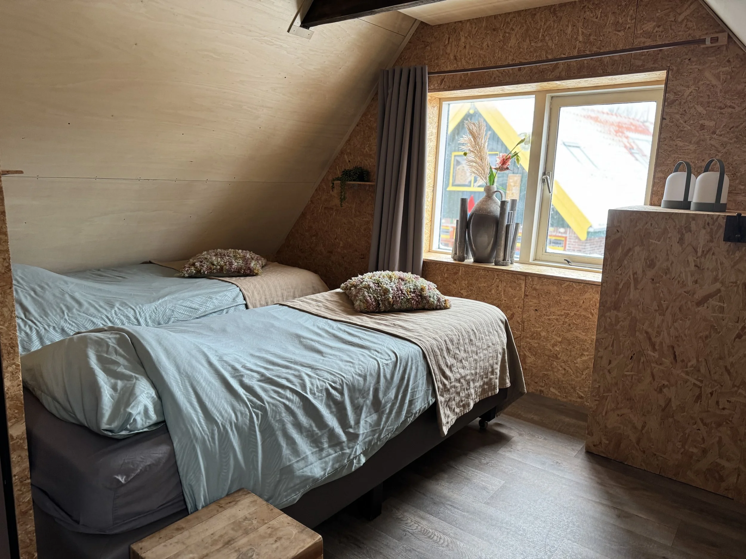 Een gezellige slaapkamer met twee eenpersoonsbedden, sierkussens, een raam met uitzicht op een kleurrijk huis buiten en een klein nachtkastje.