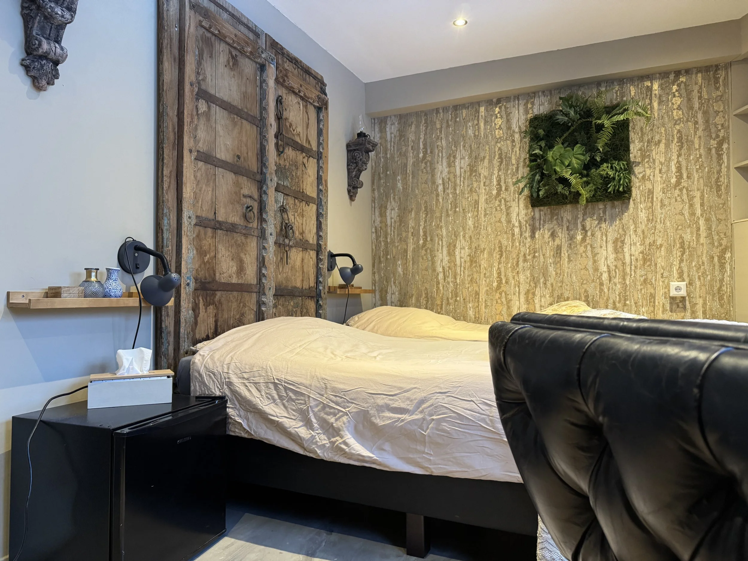 Interieur van een moderne slaapkamer met een bed met beige beddengoed, een zwarte leren fauteuil, een nachtkastje met een tissuedoosje en lampen, een grote rustieke houten schuifdeur, een groene plantenopstelling op de gestructureerde muur