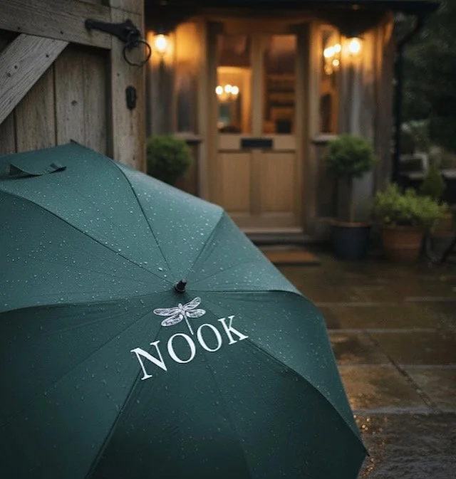 A green umbrella with the word "NOOK" and a dragonfly illustration on it, set outside a wooden house with potted plants and warm lights in the background.
