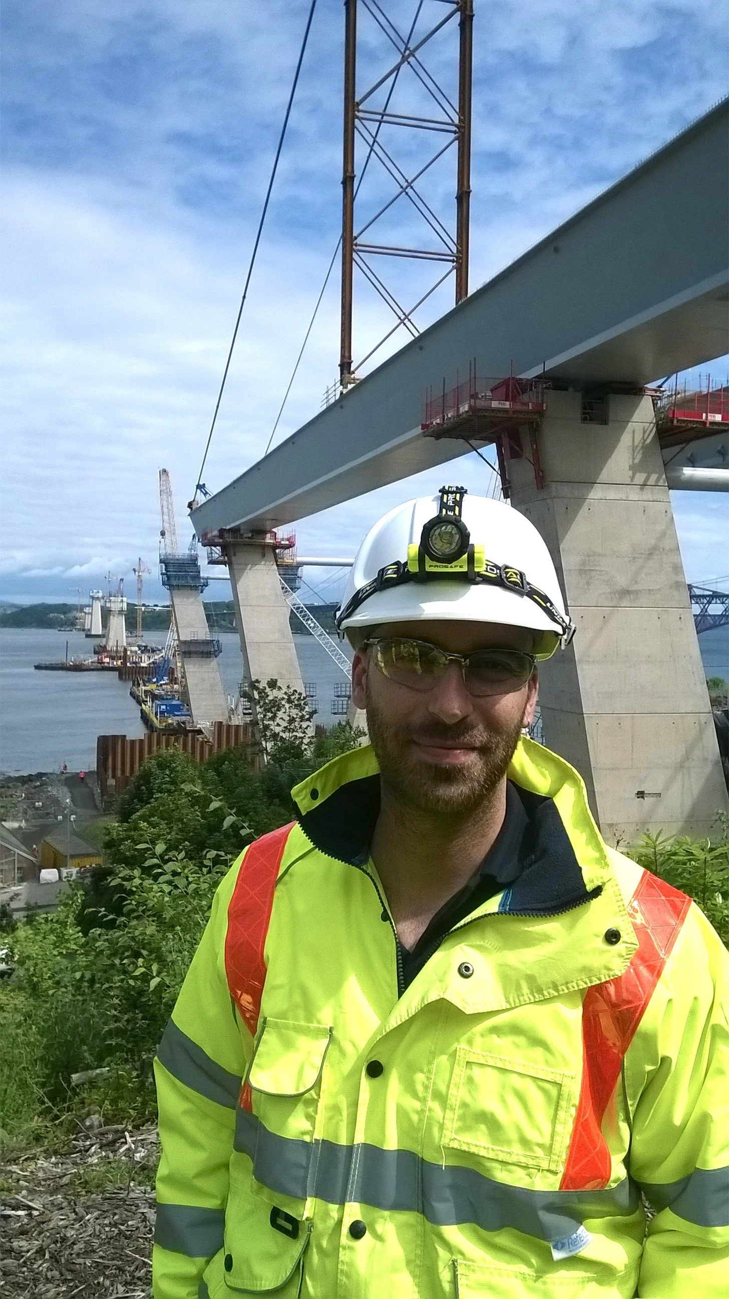A construction worker wearing a white safety helmet, safety glasses, and a bright yellow high-visibility jacket stands in front of a partially constructed bridge over water with cranes in the background.