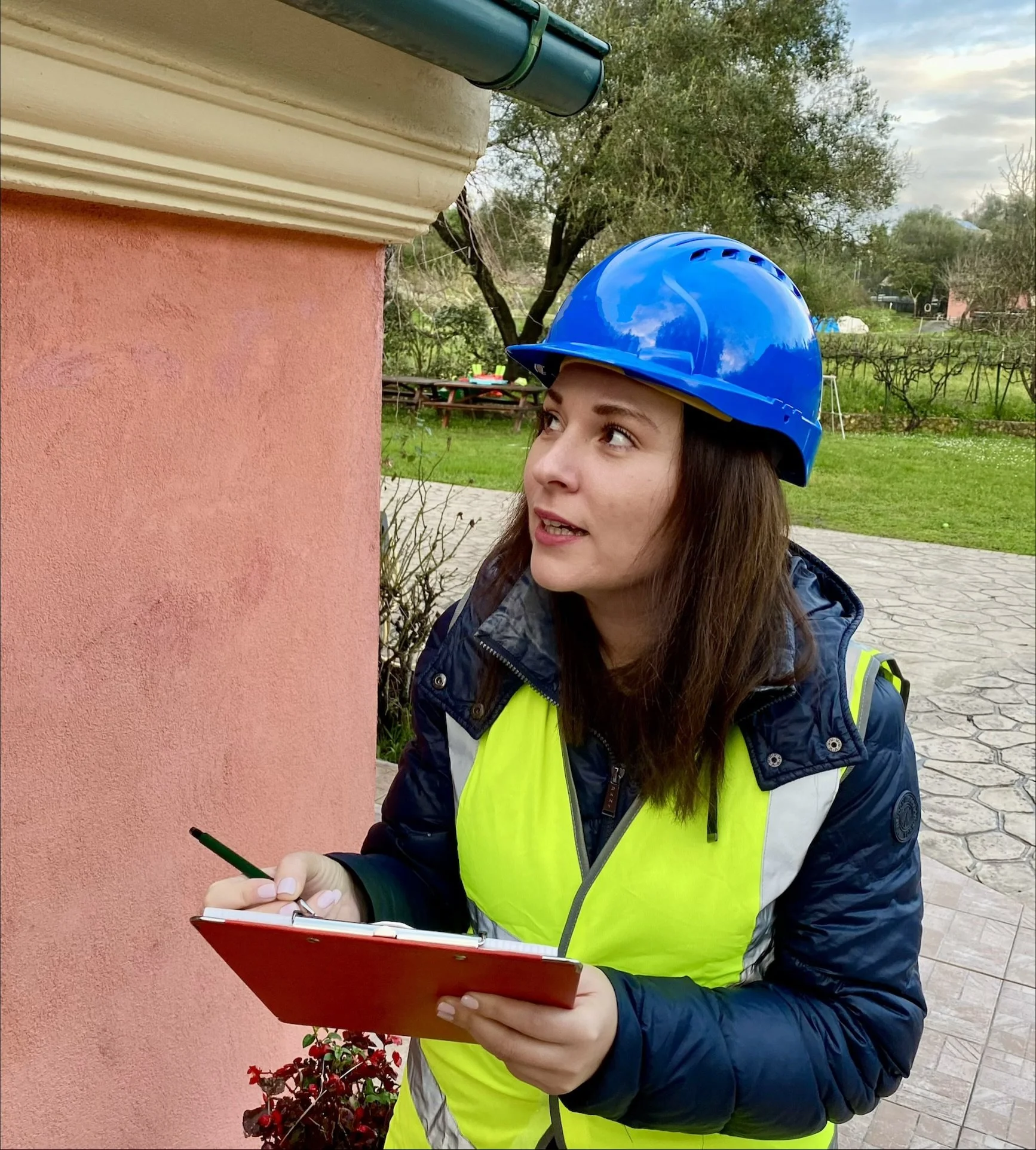 A woman wearing a blue safety helmet and reflective vest, holding a clipboard and pen, looking up at a building's corner outside.