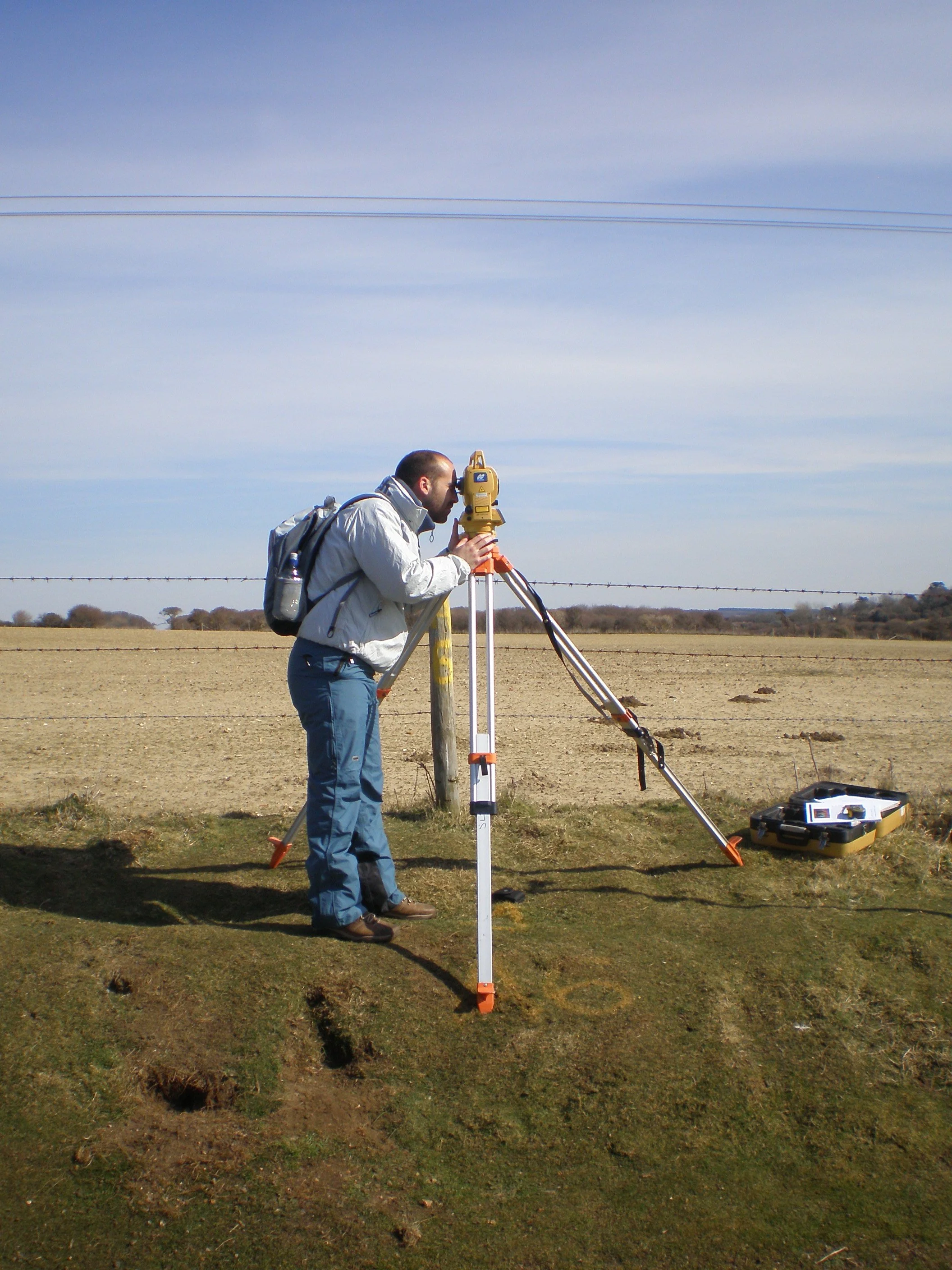 man using surveying equipment on a grassy field with a barbed wire fence and open landscape in the background