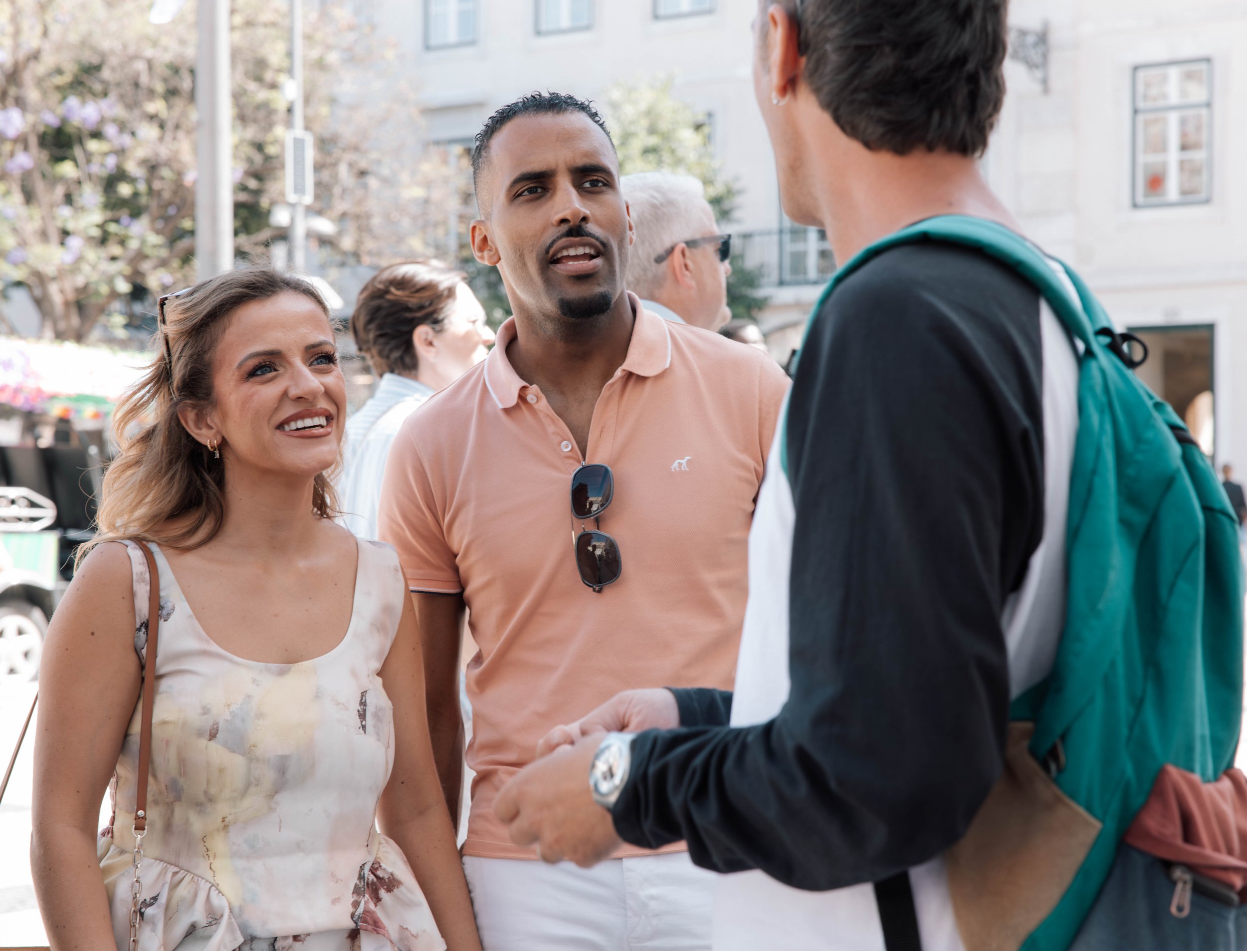 A diverse group of people engage in conversation outdoors on a city street, with buildings and trees in the background.