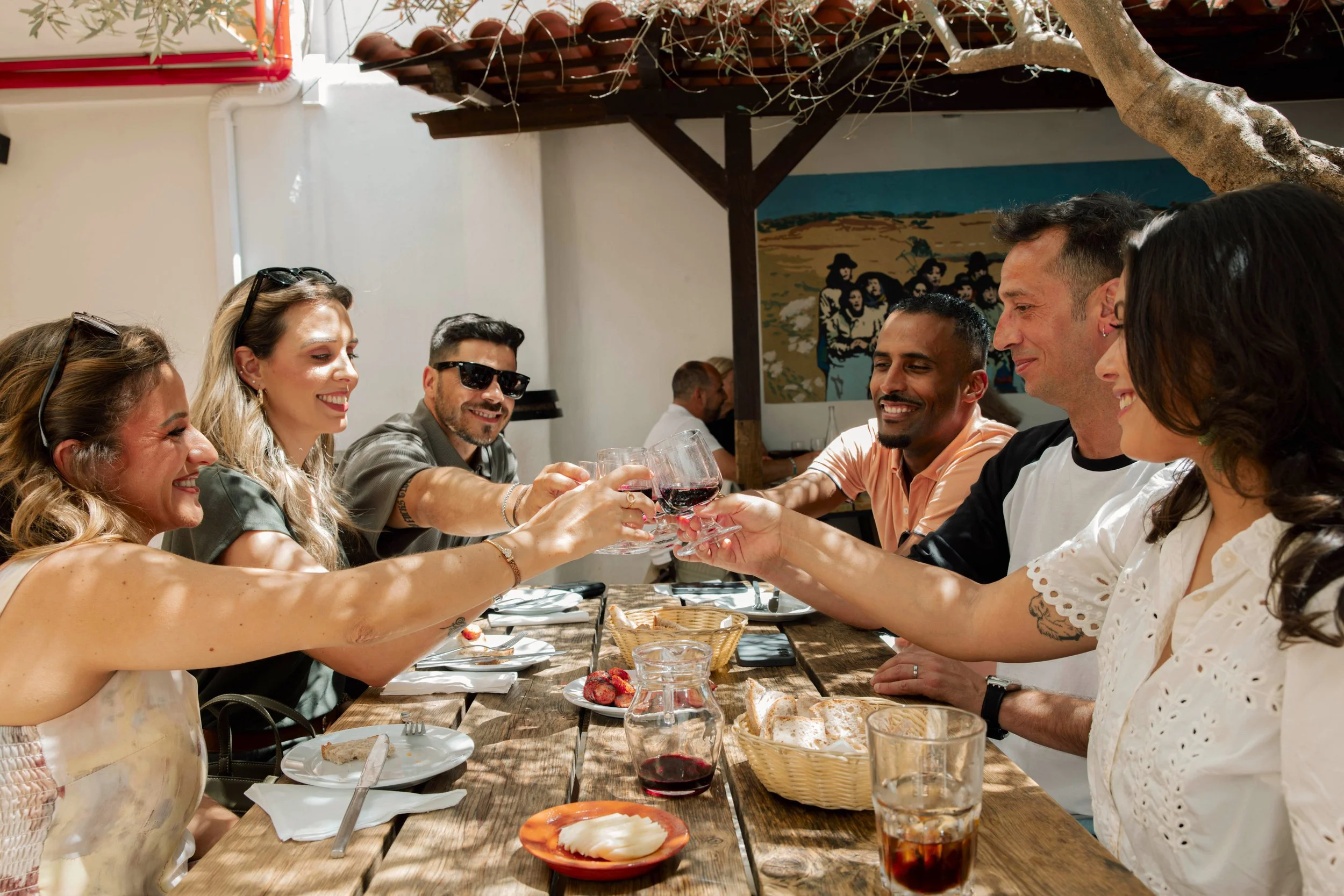 Group of six friends raising glasses of red wine for a toast at a dinner table in a bright, casual restaurant.