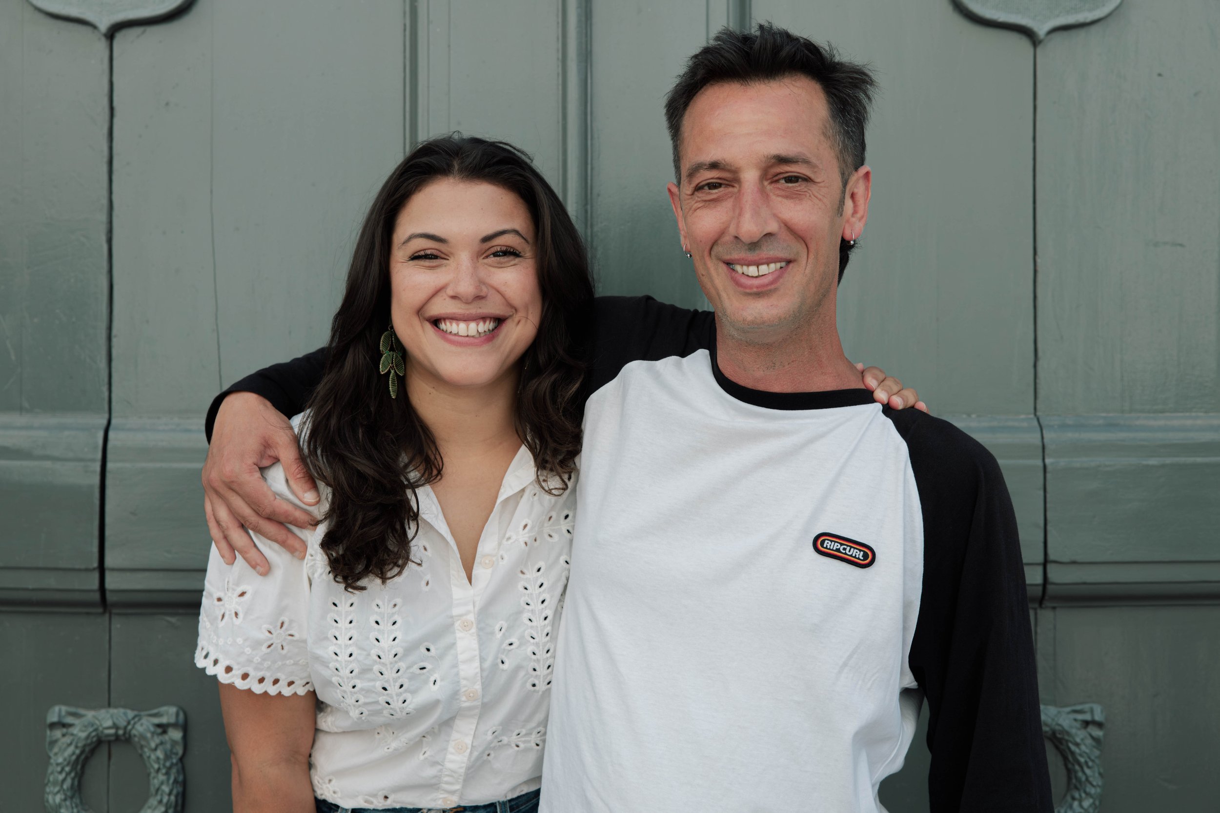 A smiling woman with dark wavy hair wearing a white eyelet blouse and green earrings stands next to a smiling man with short dark hair wearing a white and black raglan shirt with a Rip Curl logo. They are embracing each other with arms around shoulders, standing in front of a gray wooden wall.