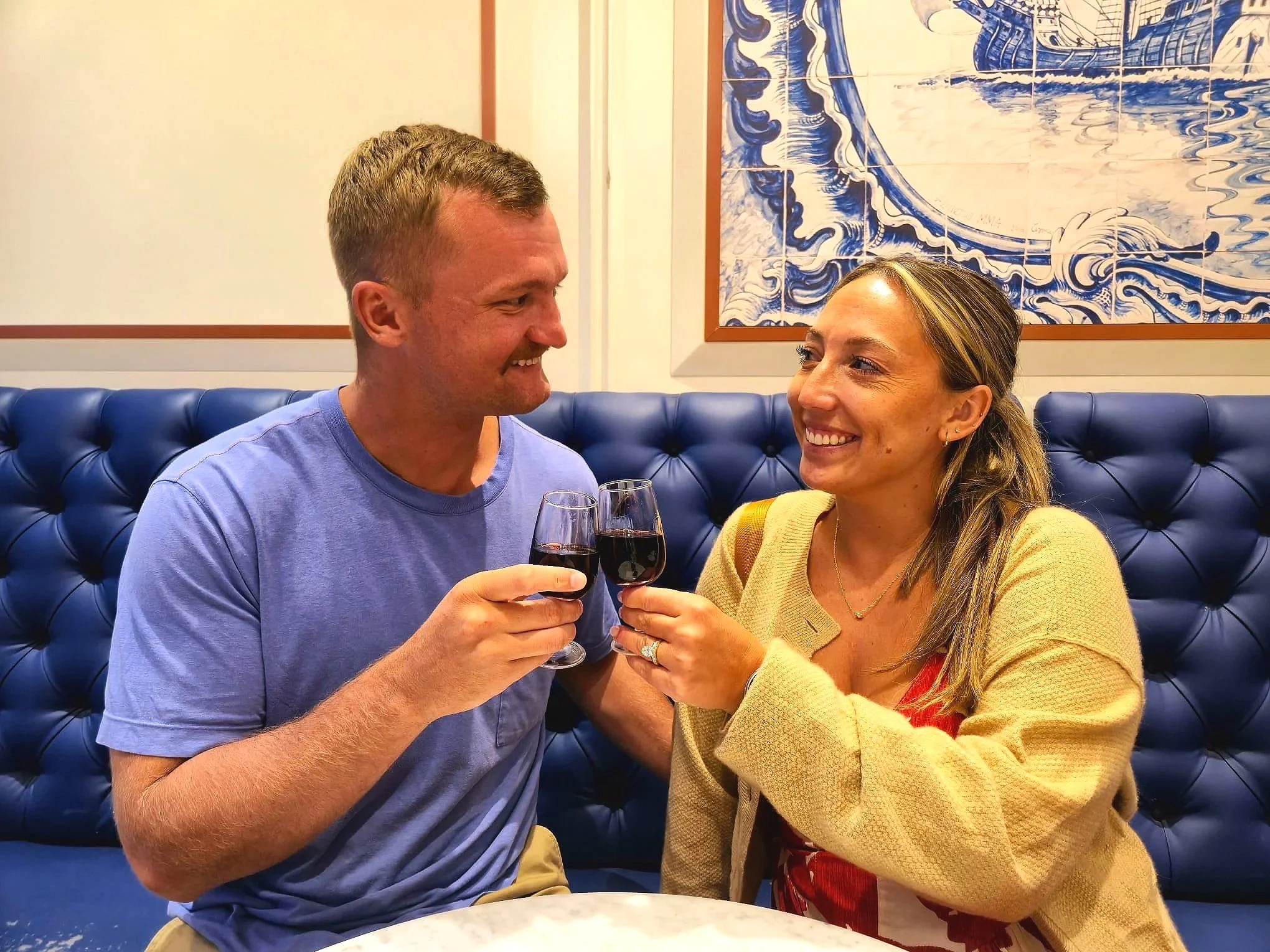 A man and a woman are sitting at a table in a restaurant, smiling at each other and clinking glasses of red wine. The man has short light brown hair and is wearing a light blue t-shirt. The woman has long hair with blonde highlights, is wearing a yellow sweater, and has multiple earrings. There is blue tufted seating and a large blue and white painting of waves on the wall behind them.