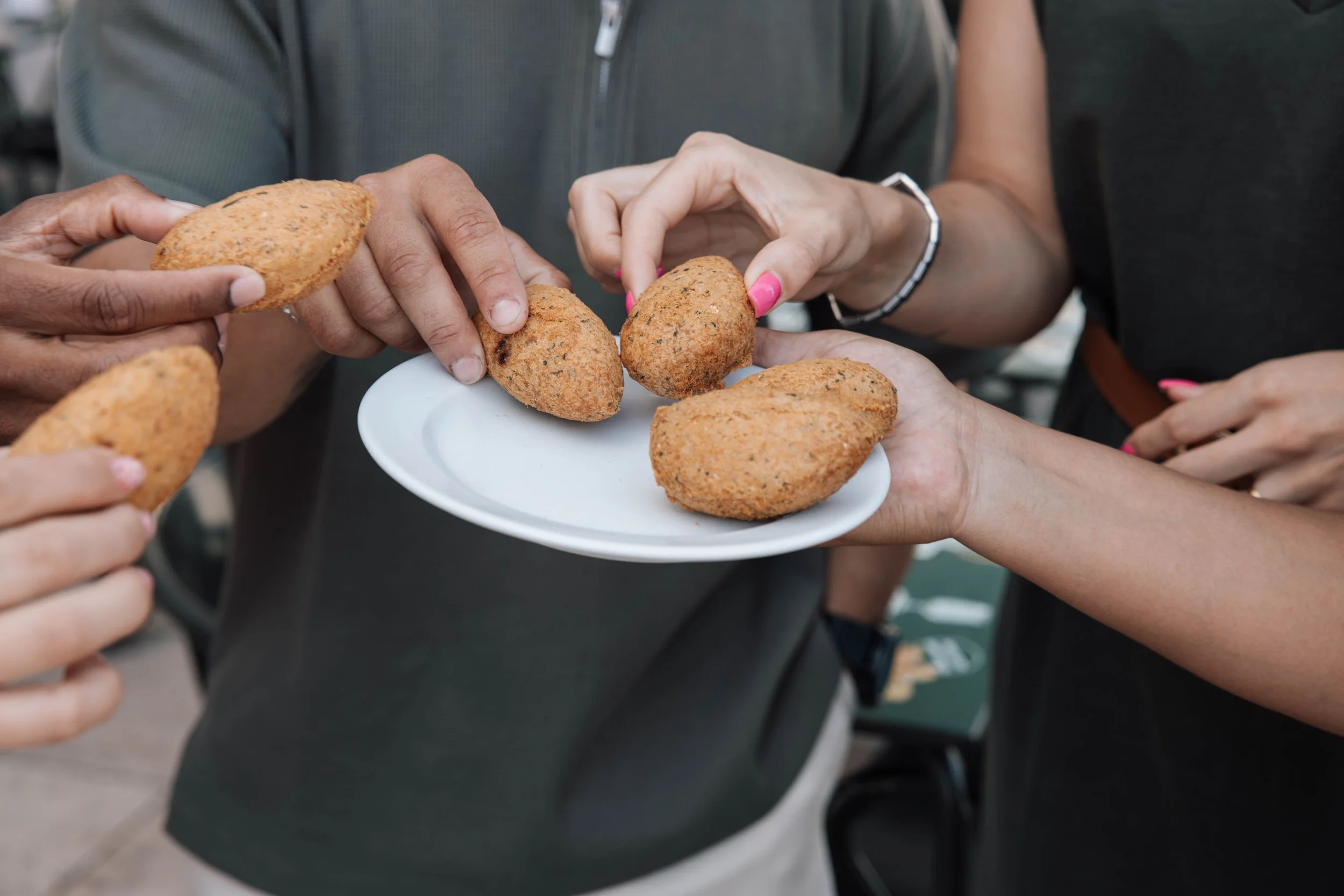Multiple hands are reaching for fried croquettes on a small white plate, with a person in the background wearing a gray shirt.