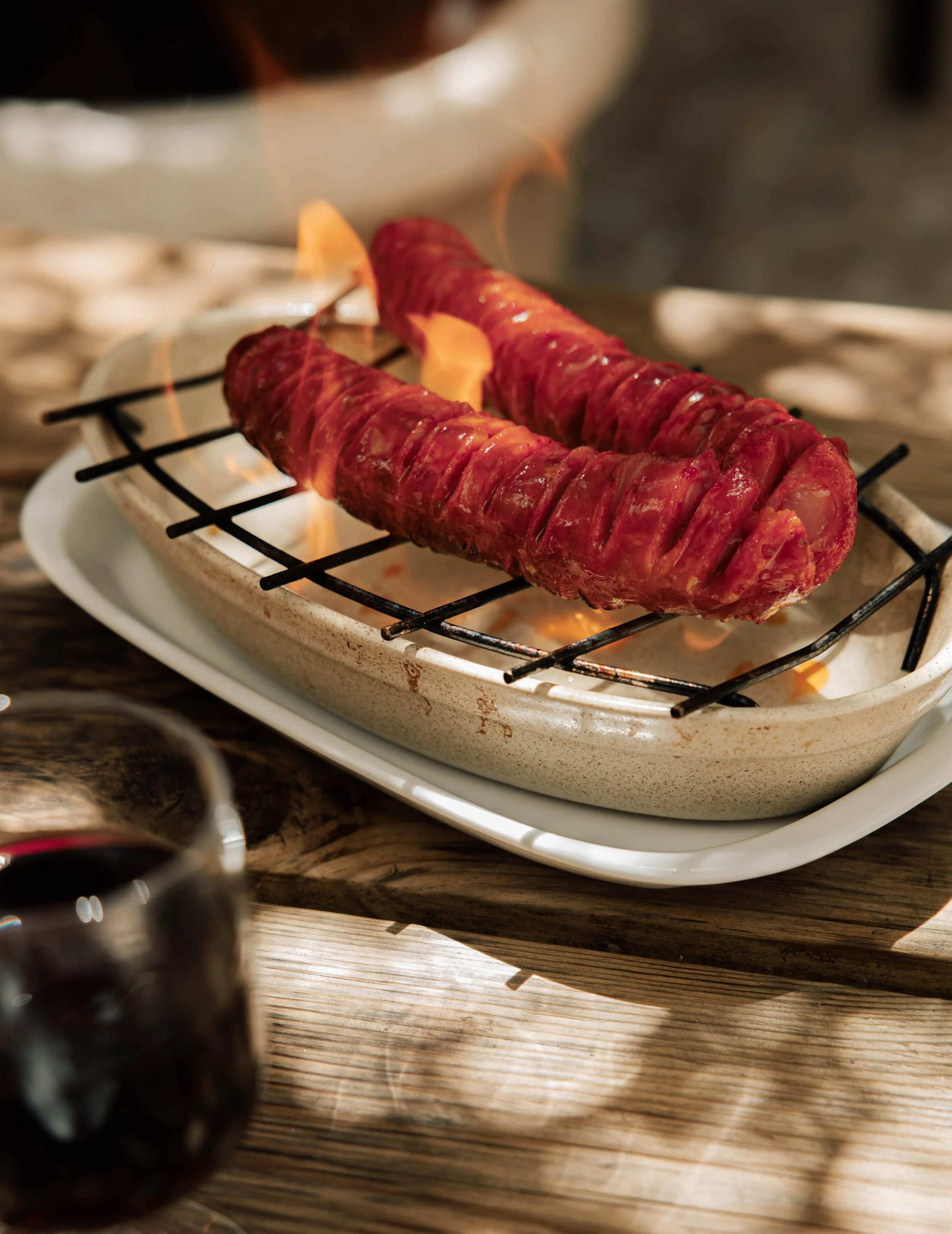 Close-up of two skewers of raw meat on a grill with flames, placed on a white plate on a wooden table in the background.