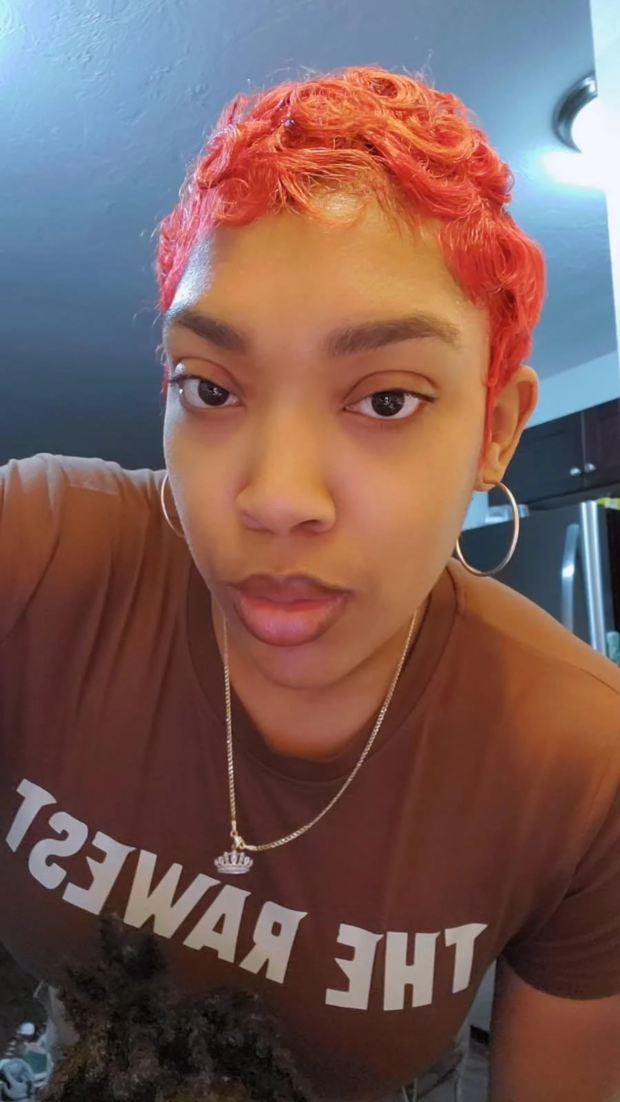 A woman with short, curly, bright red hair taking a selfie in a kitchen. She is wearing a brown T-shirt with white lettering and a silver necklace with a crown pendant.
