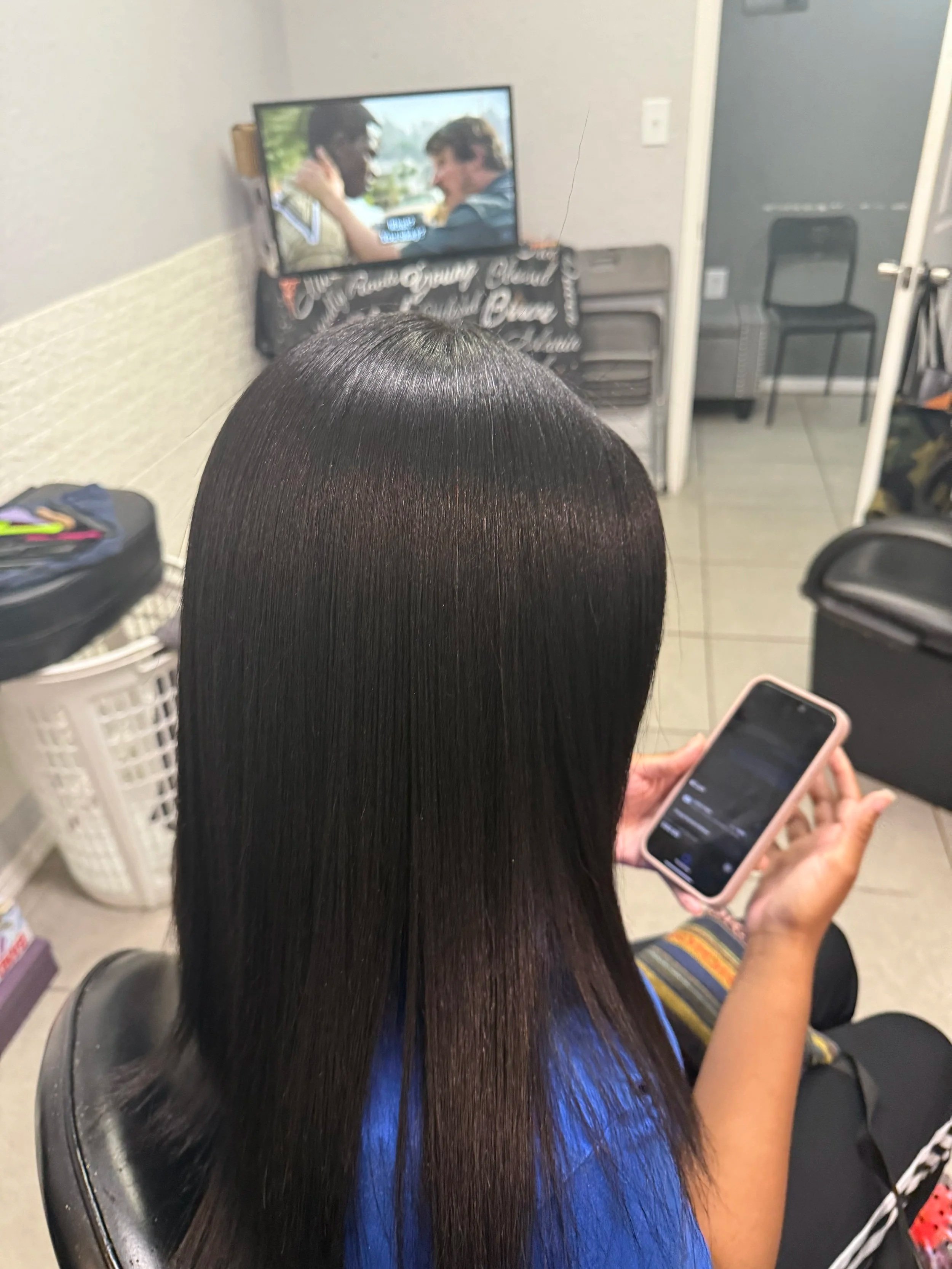 A person with long, straight, shiny black hair sitting in a salon chair, holding a smartphone, with a television on the wall in front showing a scene with two men.