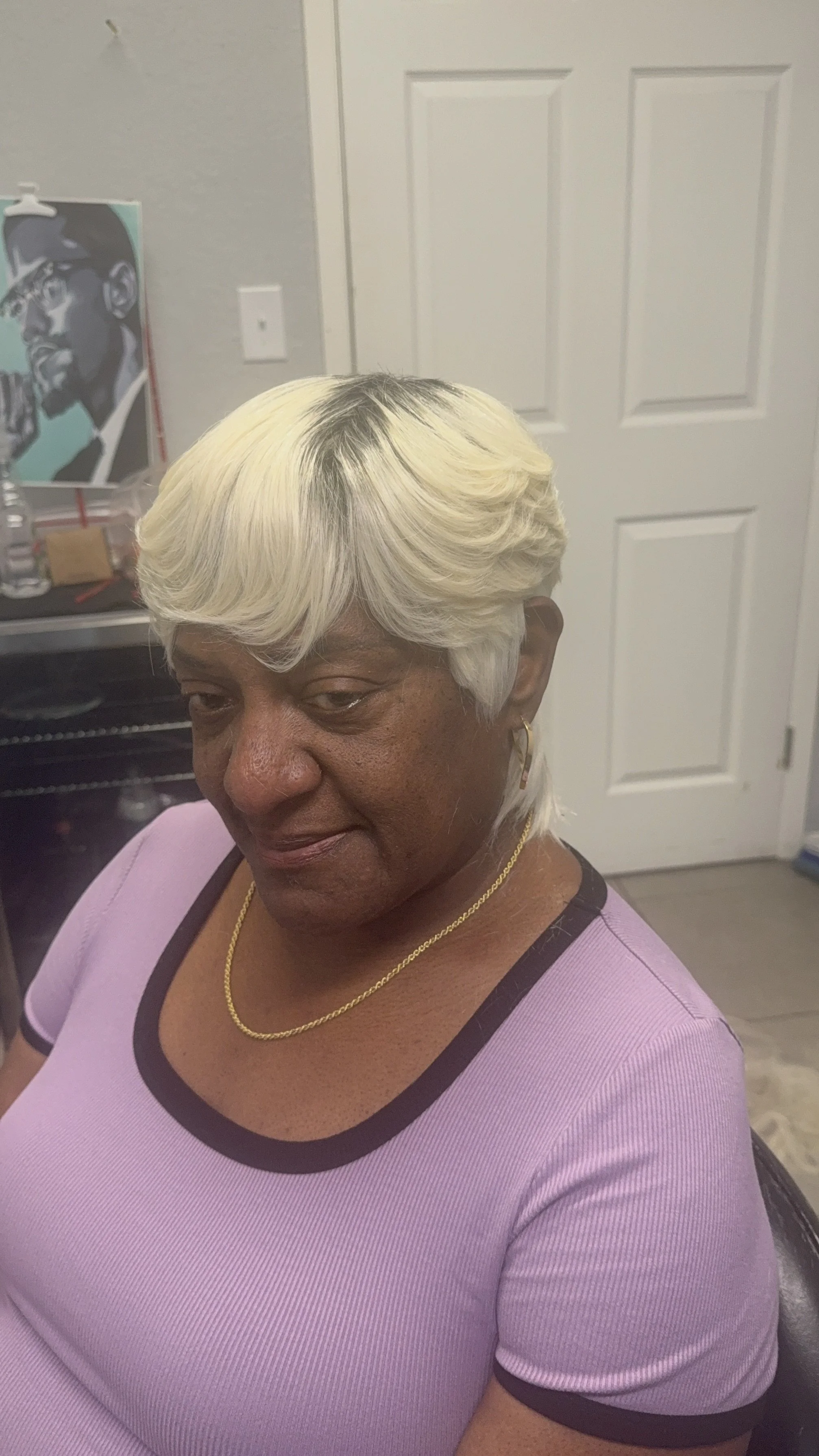 A woman with short, wavy, blonde hair, wearing a light purple shirt with dark trim, gold earrings, and a gold necklace, sitting indoors in front of a closed white door and a wall with a painting of a person wearing a hat and sunglasses.