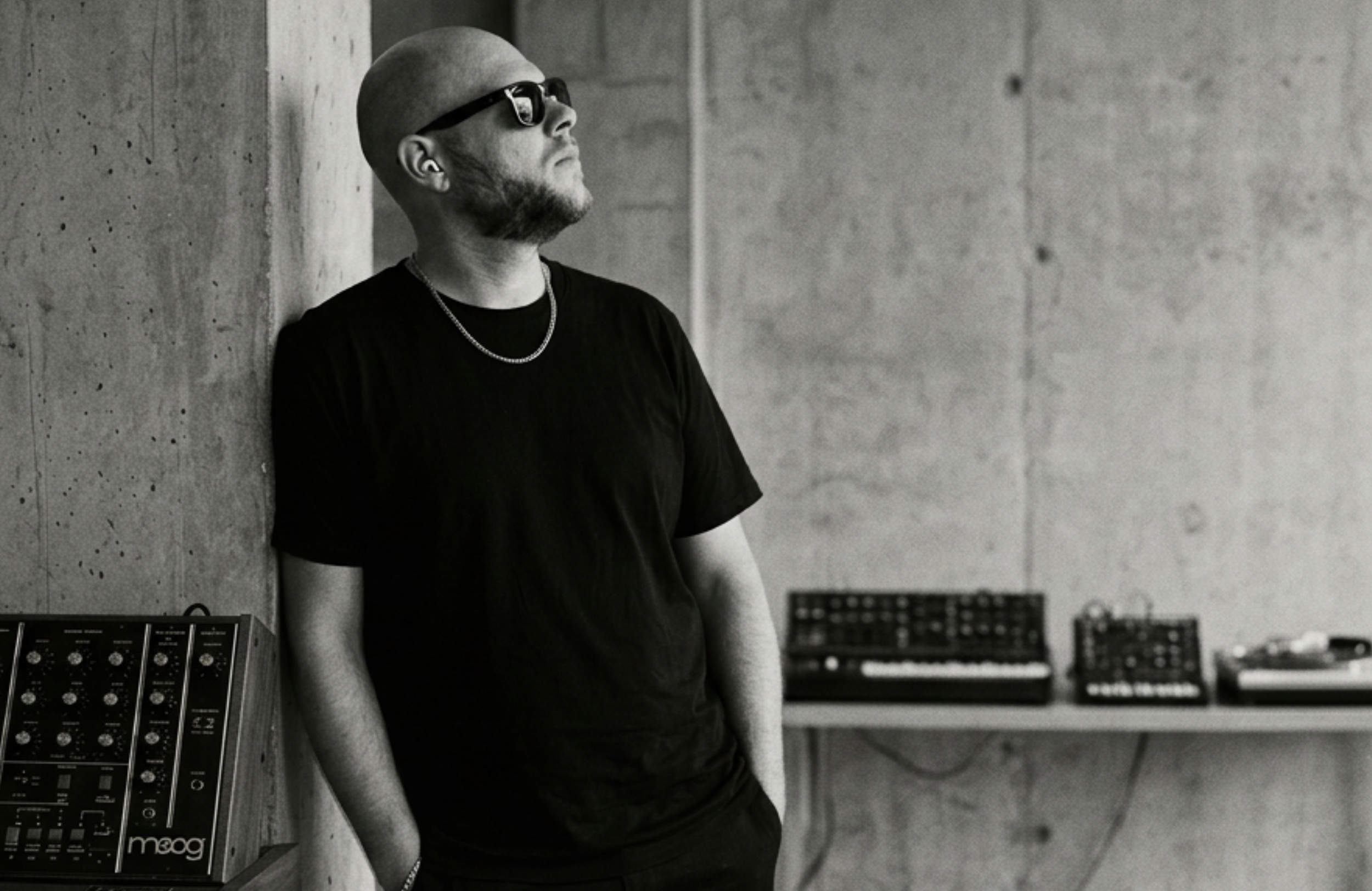 A man wearing sunglasses, a black t-shirt, and a chain necklace, leaning against a concrete wall with music equipment in the background.