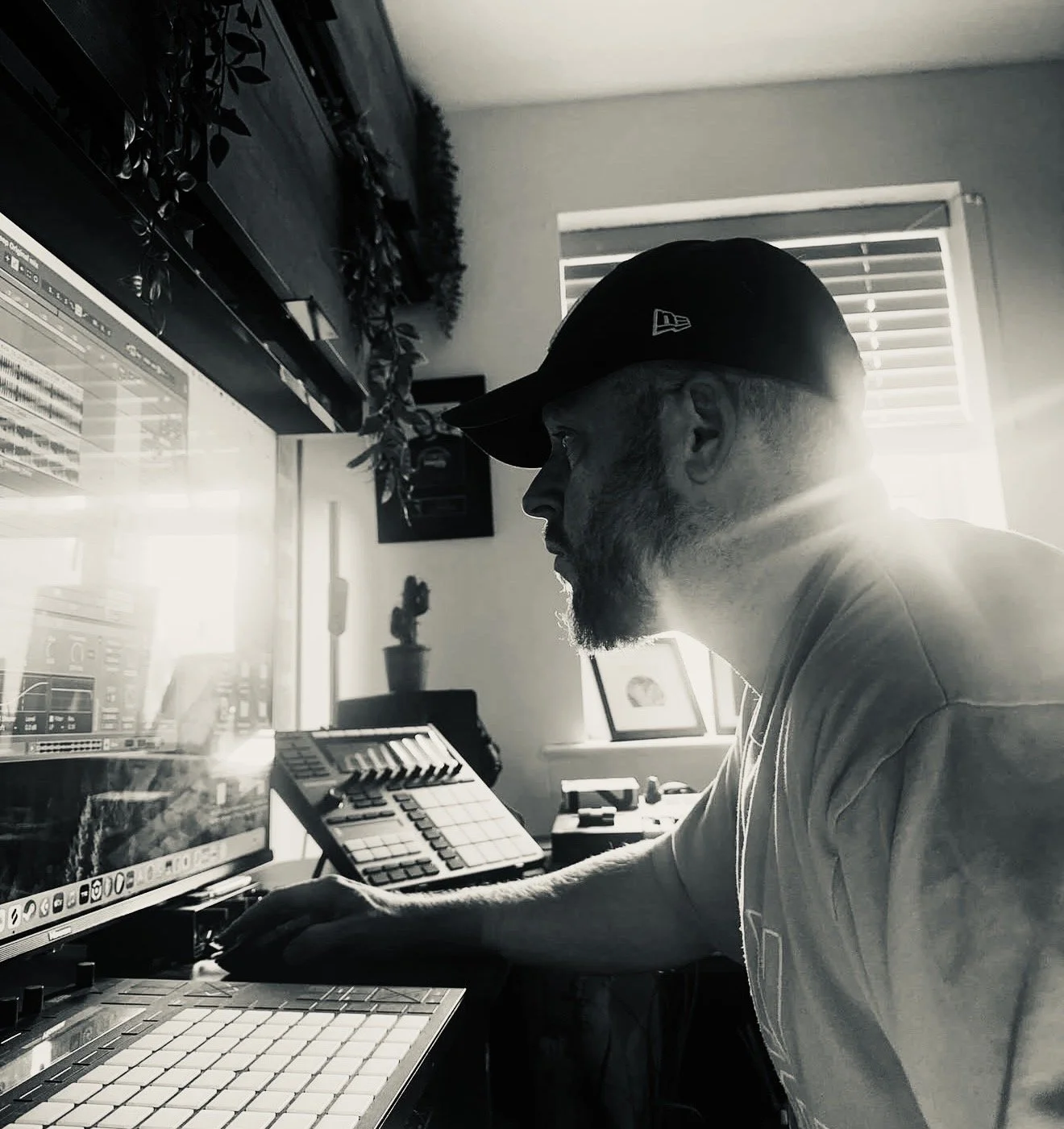 A man working on music production in a home studio, wearing a cap, in black and white with sunlight streaming through a window.