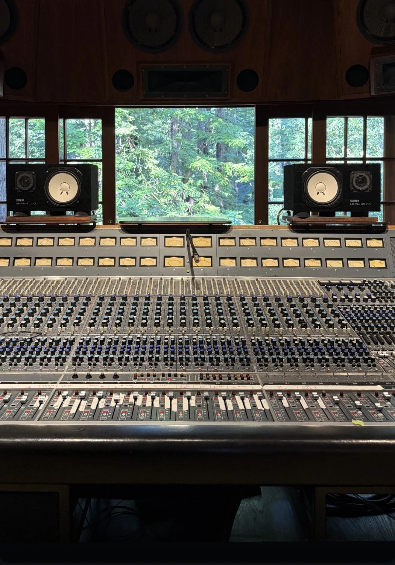 Inside a recording studio with a large mixing console, studio monitors, and a window showing a forest outside.