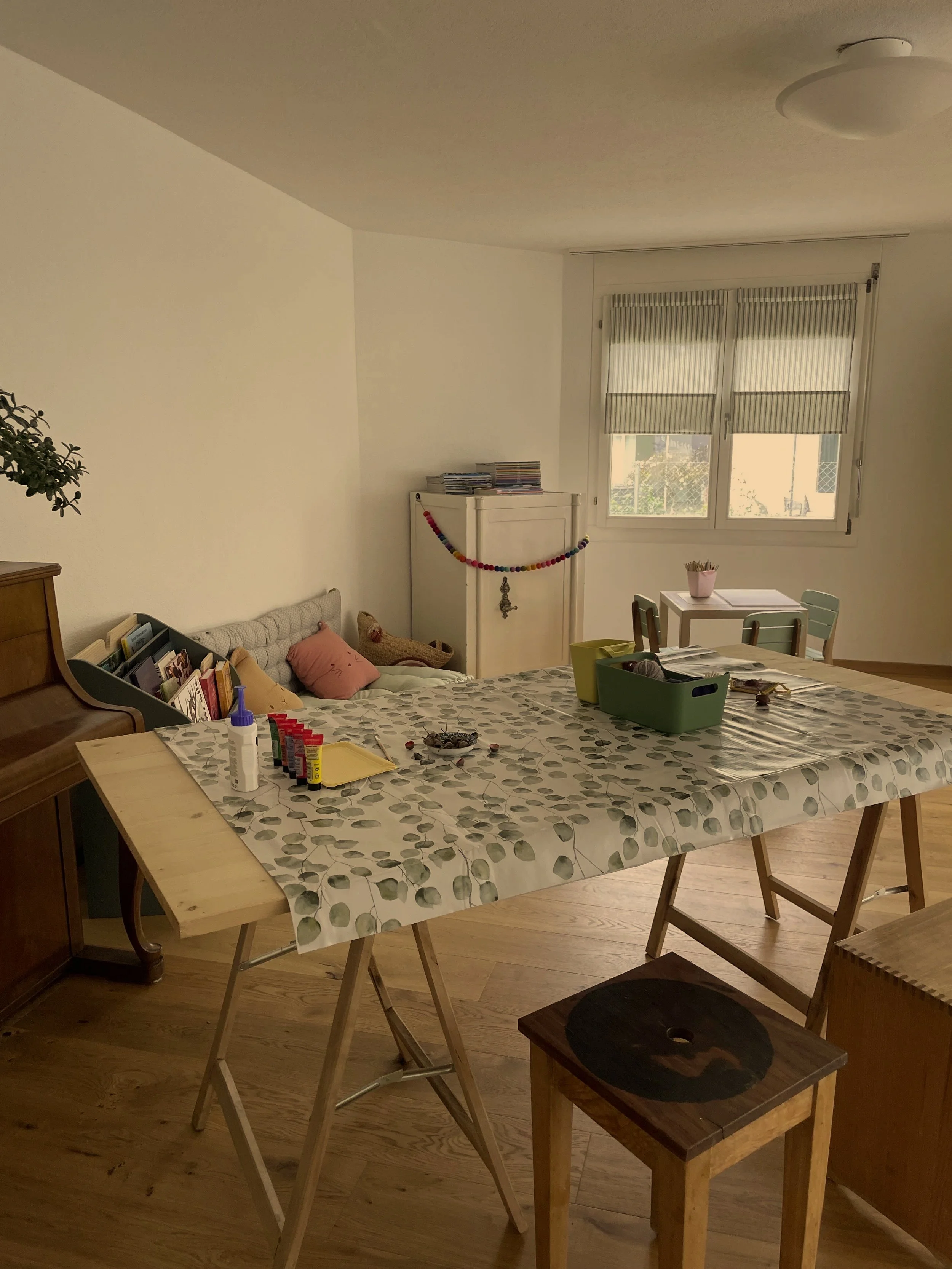 Ein Wohnzimmer mit einem großen Tisch, auf dem Bastelmaterial und Kleber liegen. Im Hintergrund eine Couch mit Kissen und eine große Fenster mit halb geöffneten Jalousien.