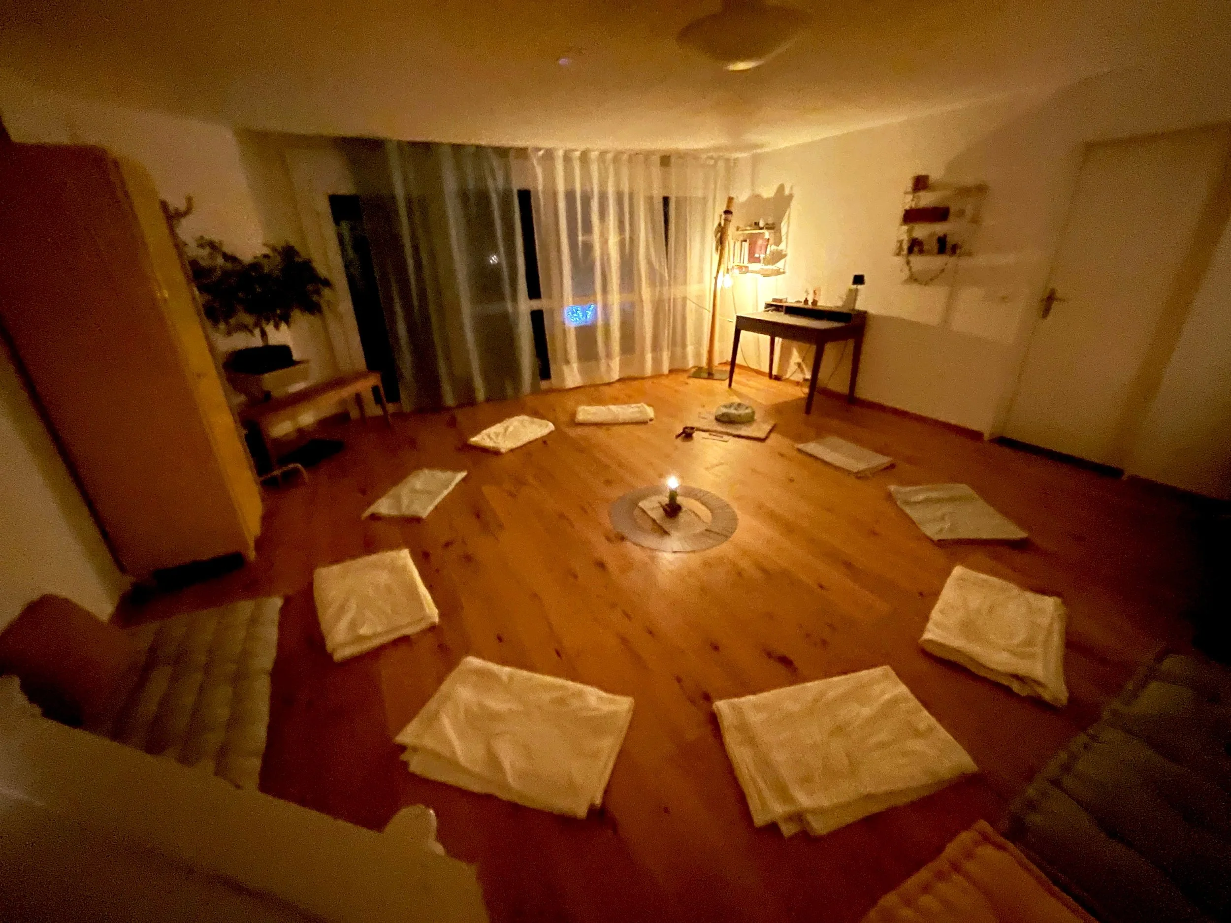 Gemütlich beleuchteter Raum mit auf dem Boden ausgelegten weißen Tüchern, einem Kerzenlicht in der Mitte und einem Fenster mit weißen Vorhängen.