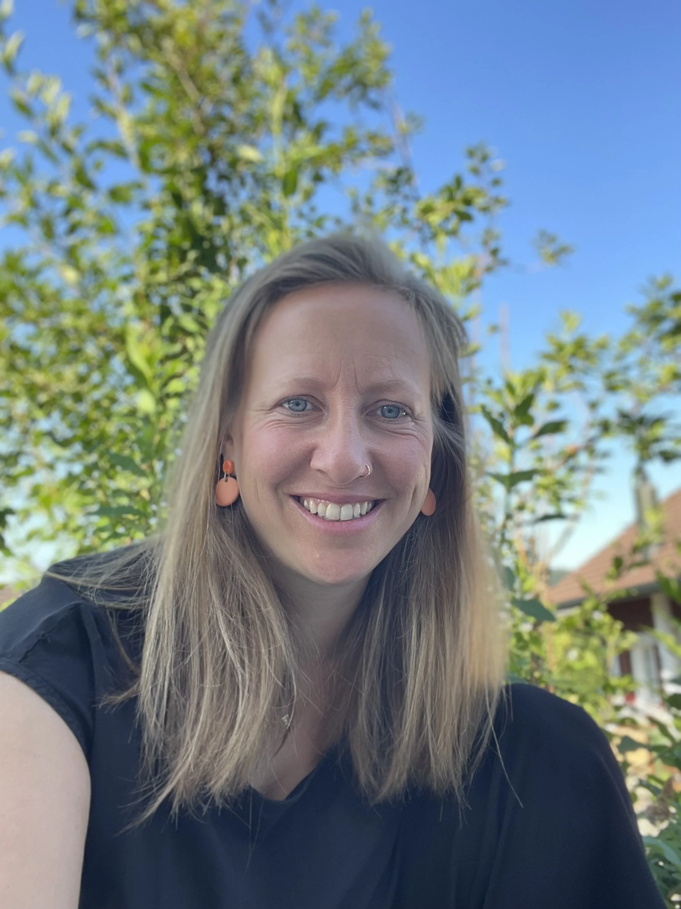 Eine lachende Frau mit langen blonden Haaren, blauen Augen, einem Nasenpiercing und orange Ohrsteckern, draußen bei blauem Himmel und grünen Bäumen im Hintergrund.