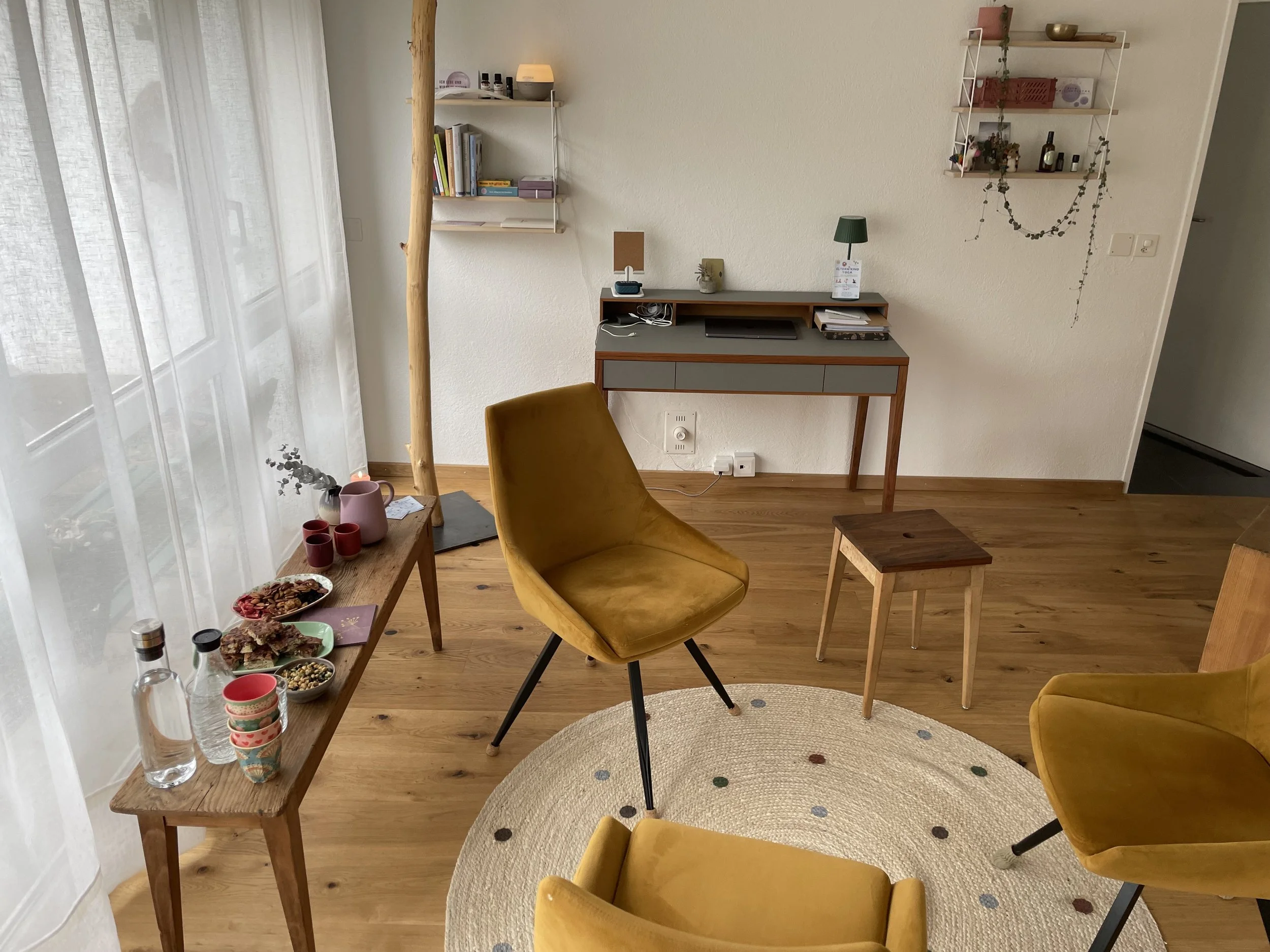 Gemütliches Wohnzimmer mit Holzfußboden, gelben Sesseln, einem kleinen Tisch, einem Schreibtisch und dekorativen Wandregalen.