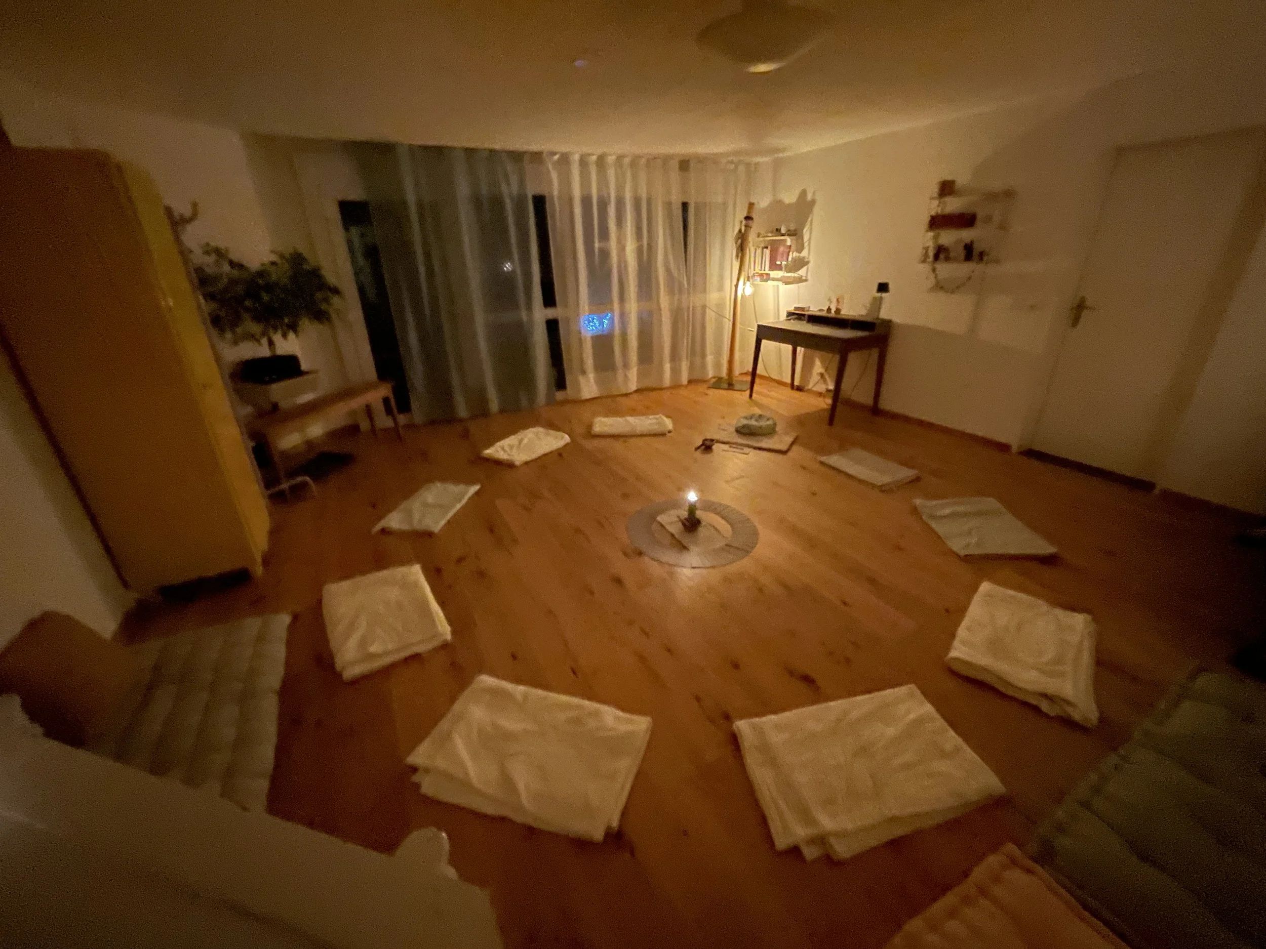 Gemütlich beleuchteter Raum mit auf dem Boden ausgelegten weißen Tüchern, einem Kerzenlicht in der Mitte und einem Fenster mit weißen Vorhängen.