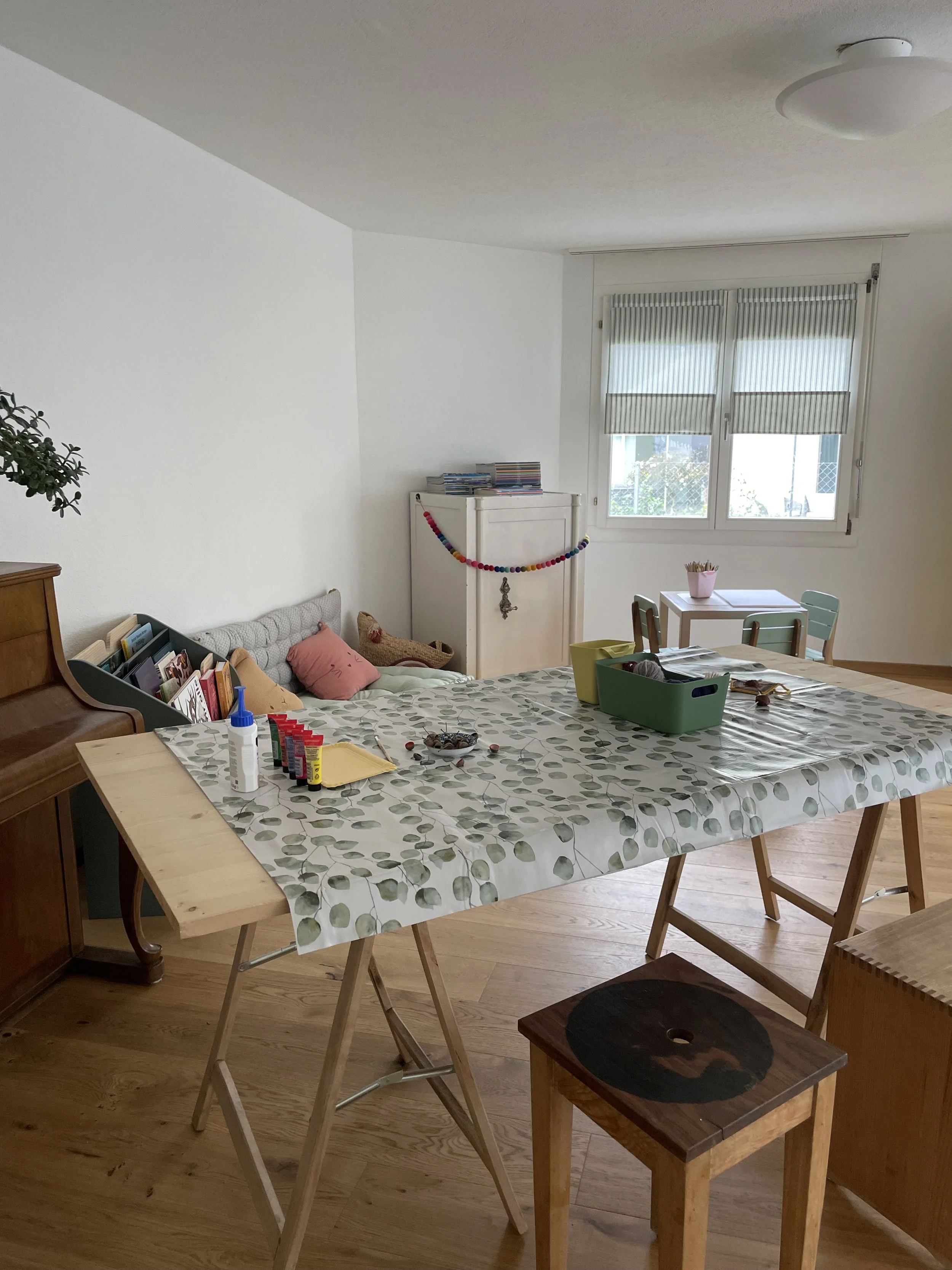 Ein Wohnzimmer mit einem großen Tisch, auf dem Bastelmaterial und Kleber liegen. Im Hintergrund eine Couch mit Kissen und eine große Fenster mit halb geöffneten Jalousien.