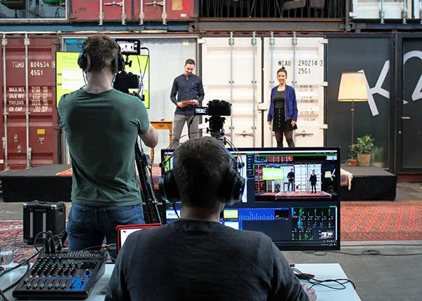 Zwei Kameraleute filmen eine Präsentation in einem Studio mit Containern im Hintergrund. Im Vordergrund sind Bildschirme und Mischpulte zu sehen. Die Szene wirkt industriell und technisch.