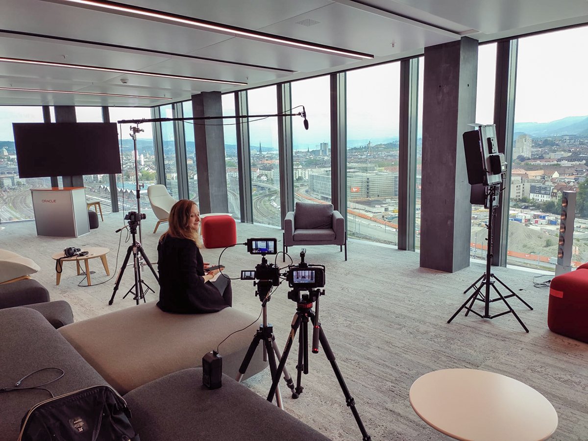 Raum mit moderner Einrichtung und großem Fenster, das eine städtische Aussicht bietet. Kameraausrüstung und Beleuchtung sind eingerichtet, mit einer Person, die auf ein Sofa schaut. Ein Bildschirm auf einem Podium im Hintergrund mit der Aufschrift 'O