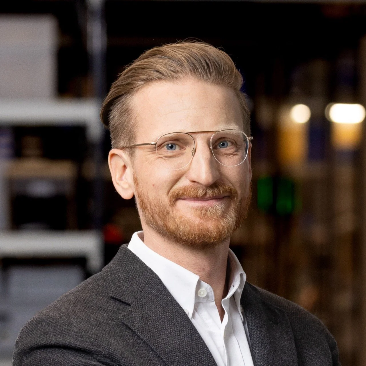 Mann mit Brille und rotem Bart in einem Anzug, lächelt leicht, unscharfer Hintergrund.