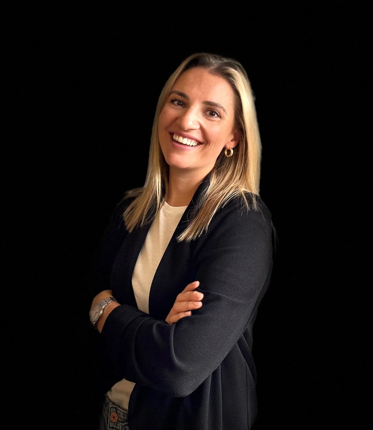 Lächelnde Frau mit blondem Haar vor schwarzem Hintergrund, gekleidet in schwarzer Jacke und weißem Oberteil, mit verschränkten Armen.
