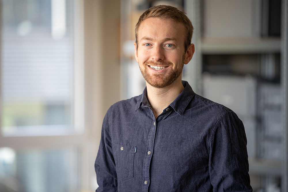 Ein lächelnder Mann in einem dunkelblauen Hemd steht in einem hellen Raum. Im Hintergrund sind Regale und ein Fenster zu sehen.