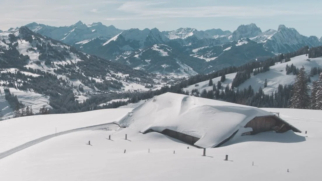 Verschneite Berghütte in den Alpen, umgeben von Bergen und Wäldern.