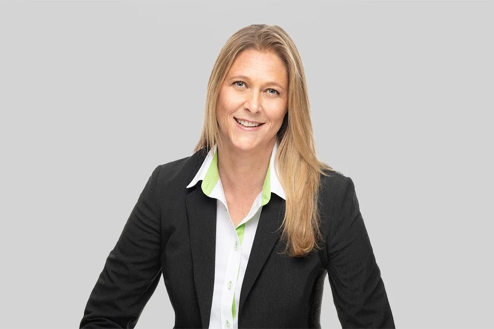 Frau in einem schwarzen Anzug mit weißem Hemd, vor grauem Hintergrund, lächelnd.