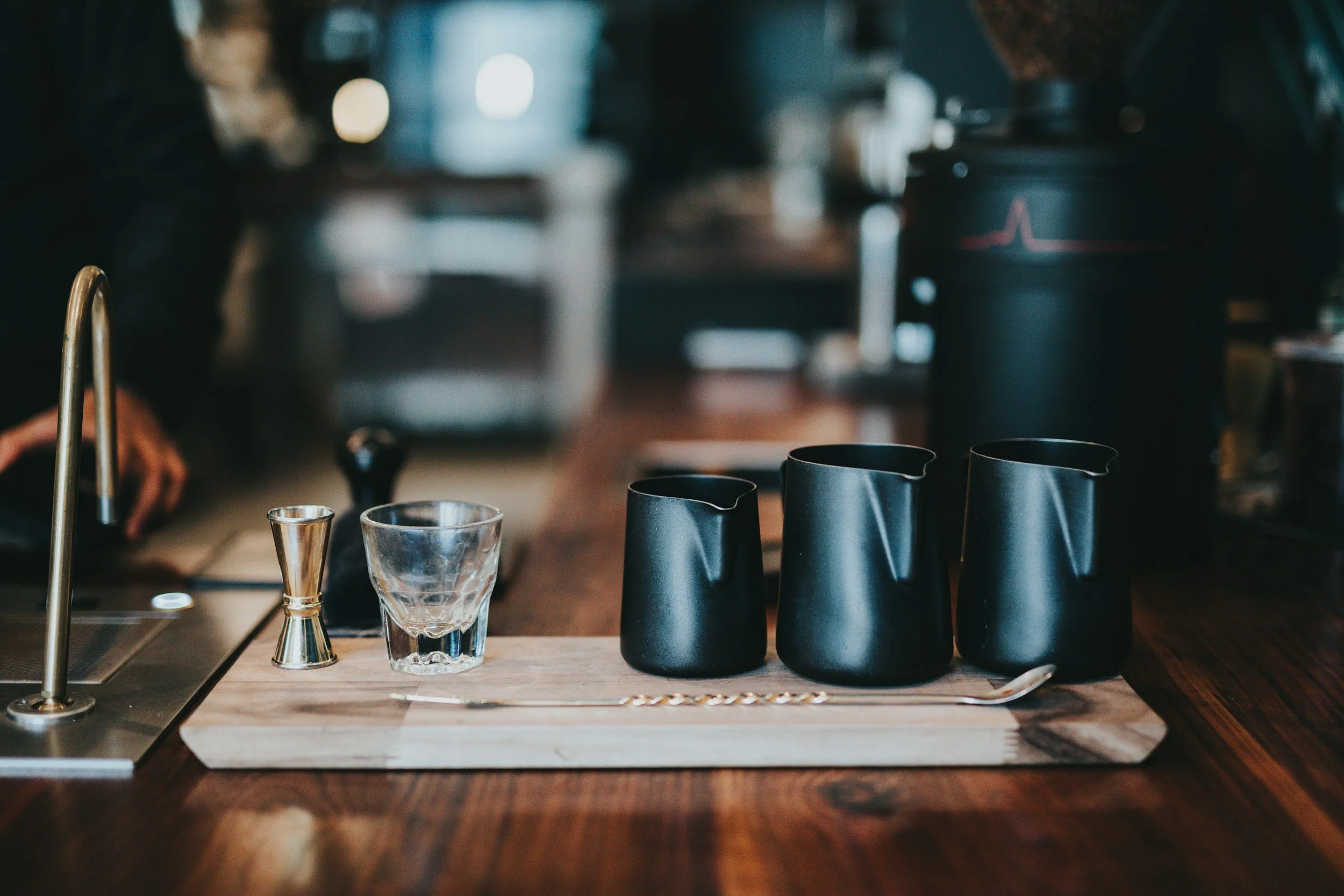 Black coffee pitchers, a glass, and a metal jigger on a wooden tray at a coffee shop counter.