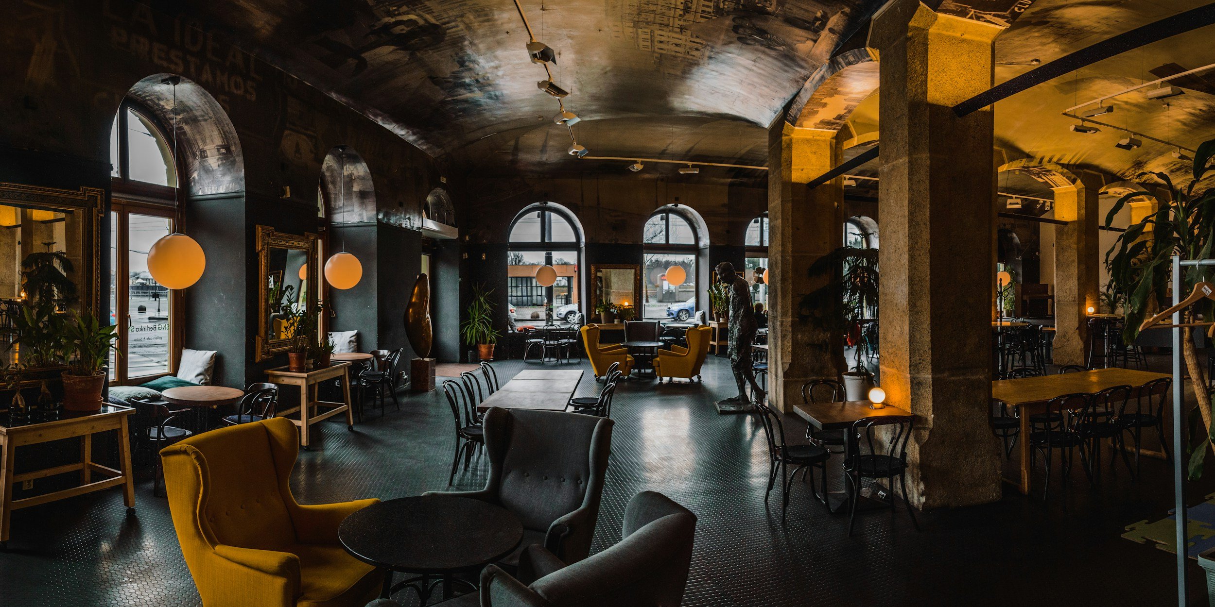 Interior of a trendy cafe with large arched windows, various seating arrangements including armchairs and tables, plants, and modern decor with warm lighting.